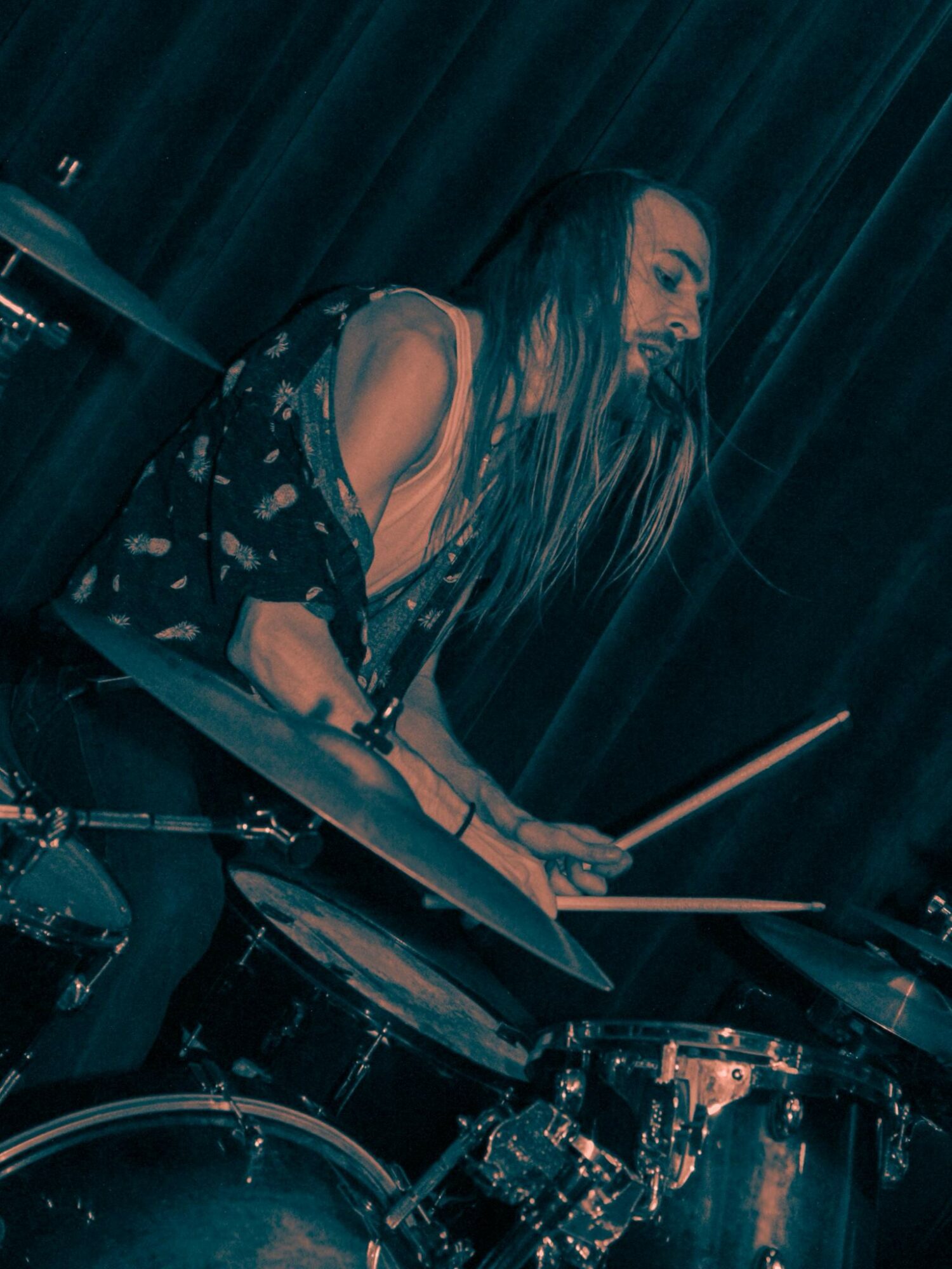 Woman playing drums with long hair, dark background, wearing a patterned top, focused on her performance.