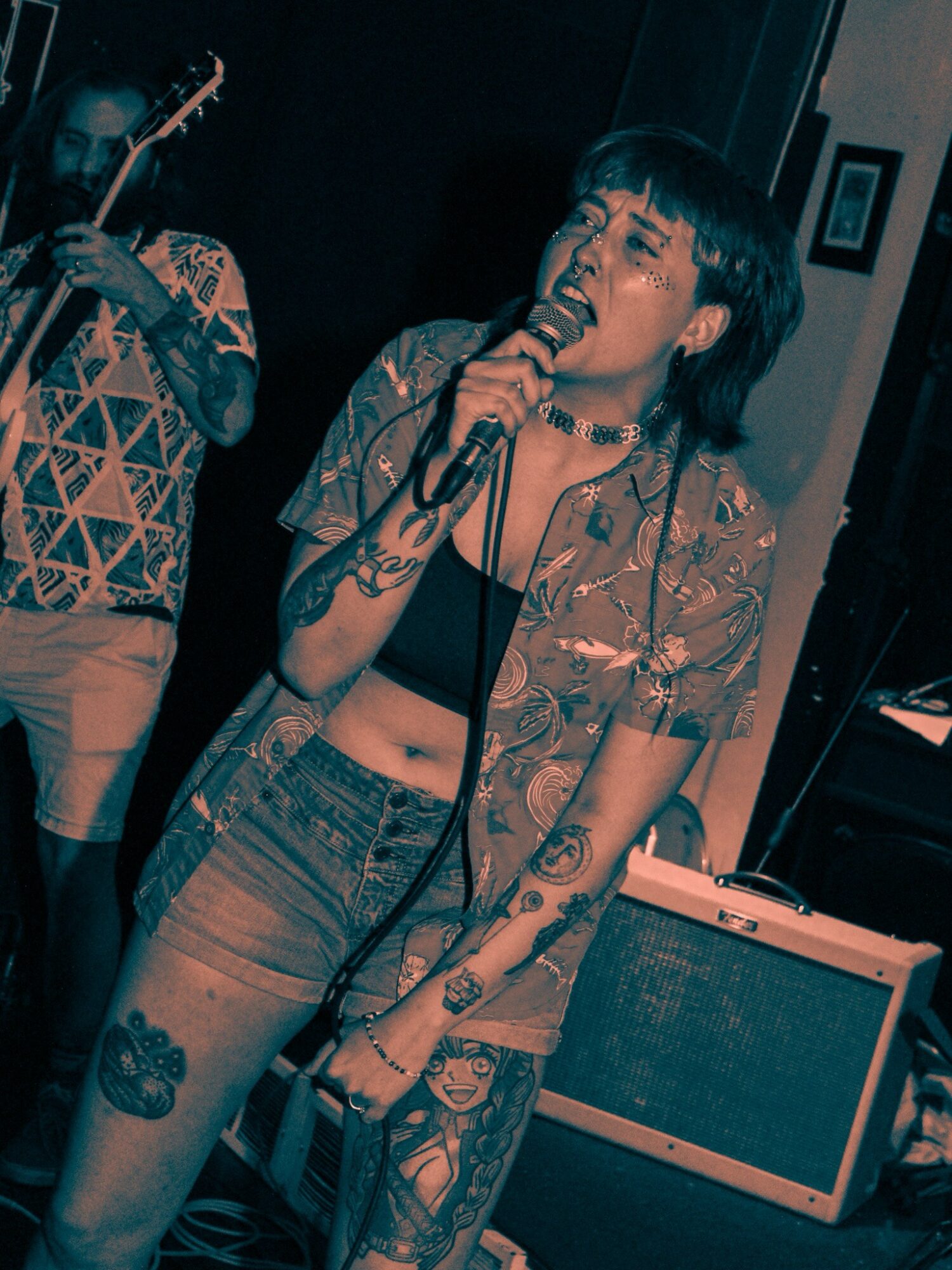 Person singing into a microphone with tattoos, wearing a patterned shirt and jeans, in a dimly lit room.