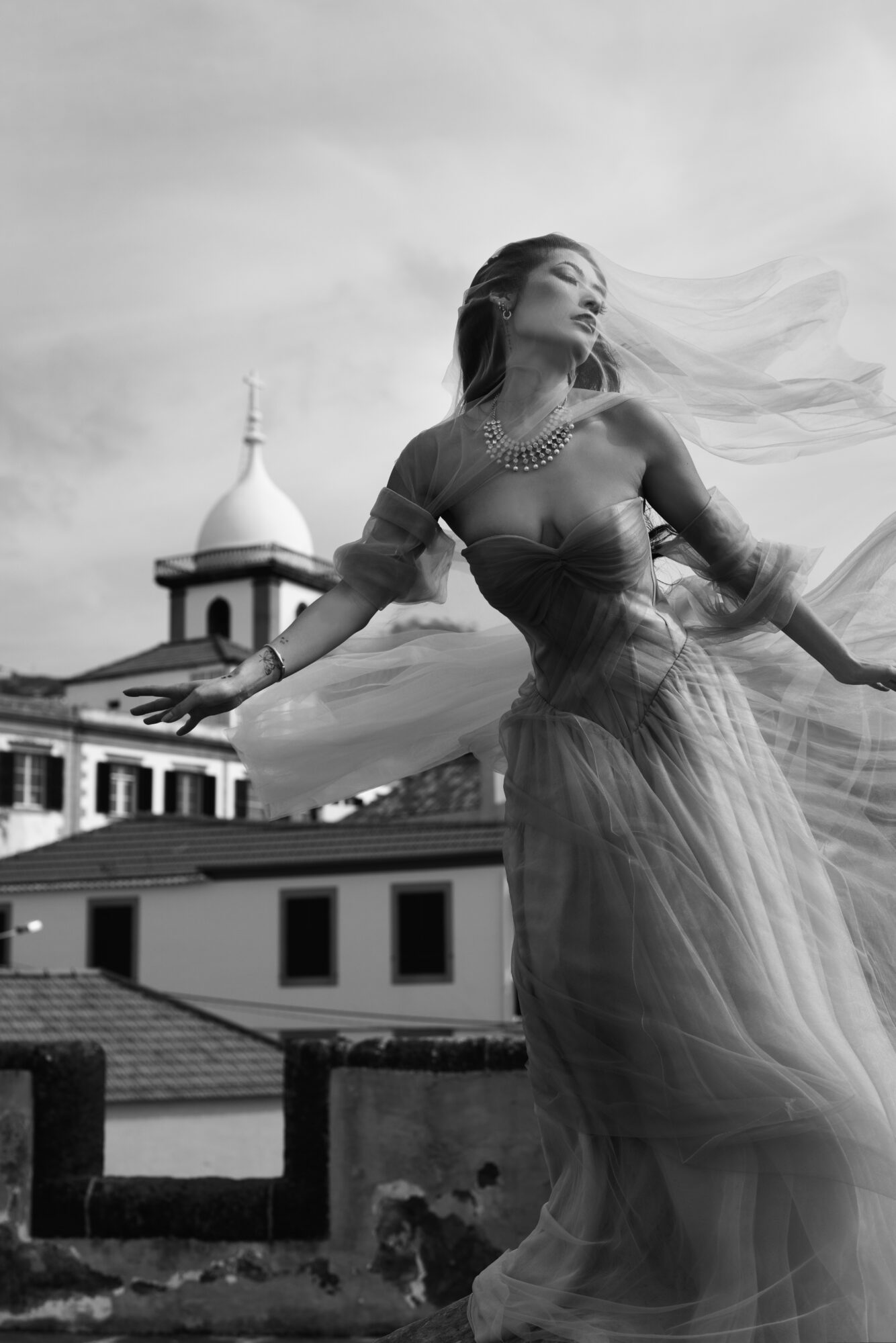 A woman in a flowing dress stands outdoors with her arms outstretched, against a background of buildings and a dome, in black and white.