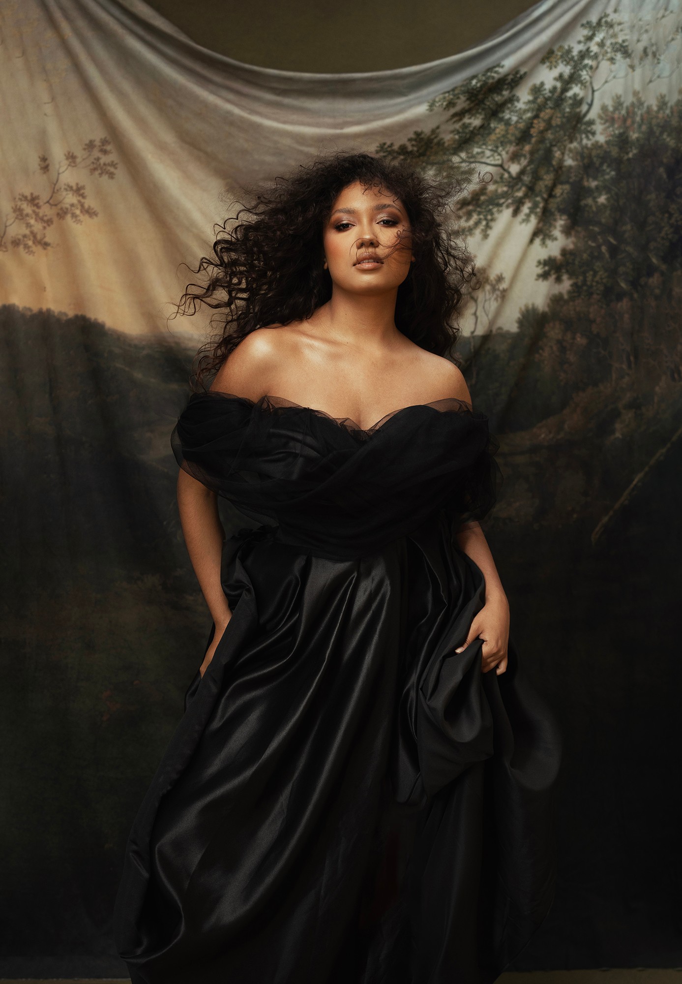 Woman with curly hair in a black off-shoulder dress standing in front of a nature-themed backdrop