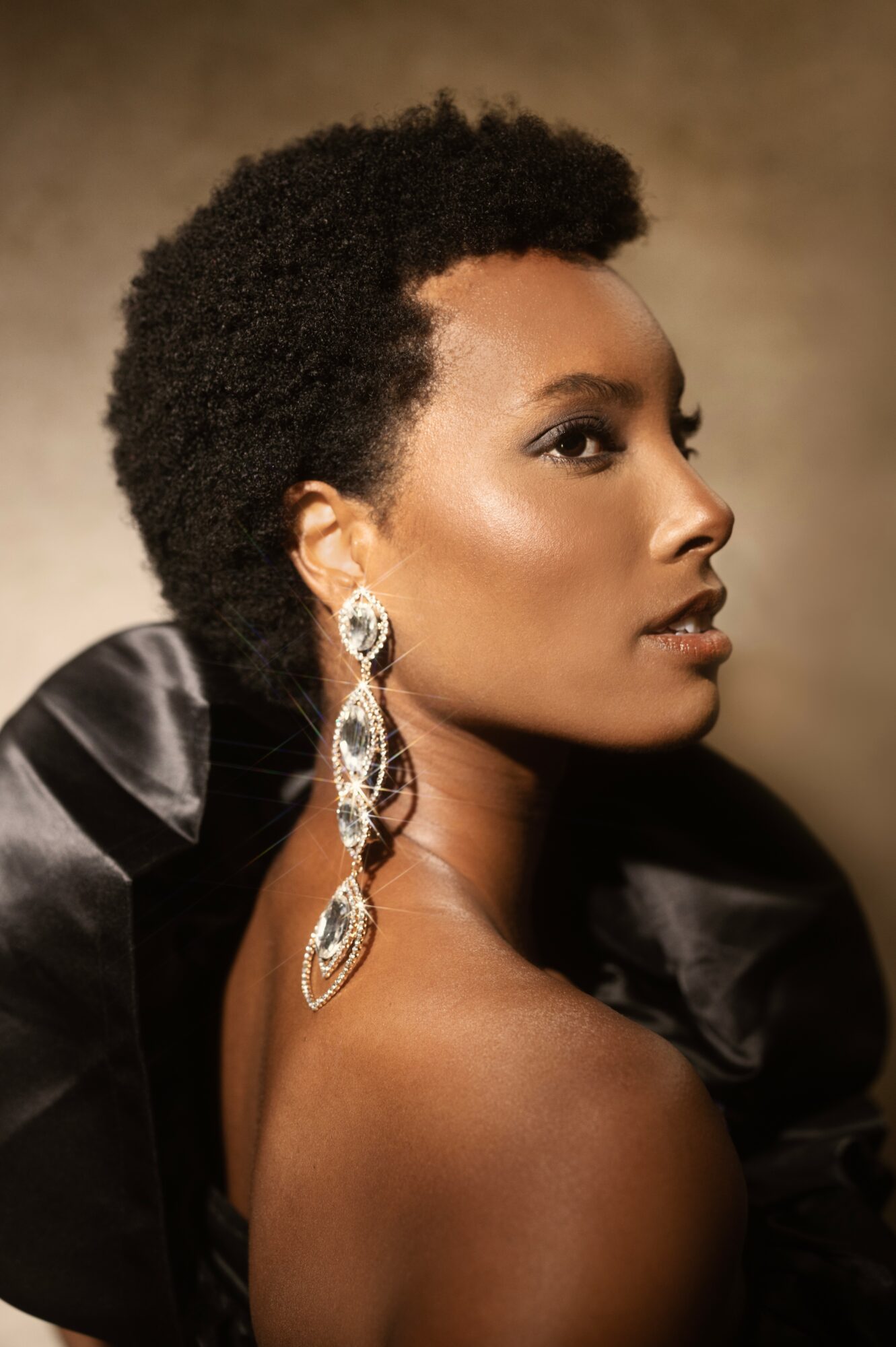 Profile of a woman with short curly hair, wearing large earrings and a black satin garment, against a neutral background.