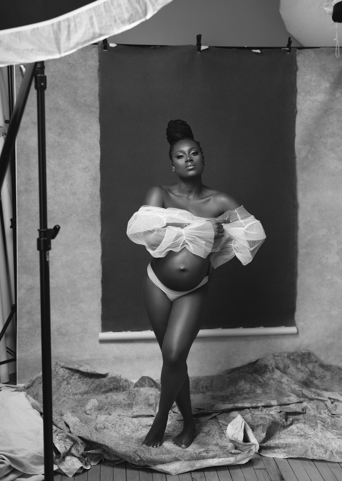 A woman stands in front of a dark backdrop, wearing an off-shoulder top and underwear, posing with arms crossed.