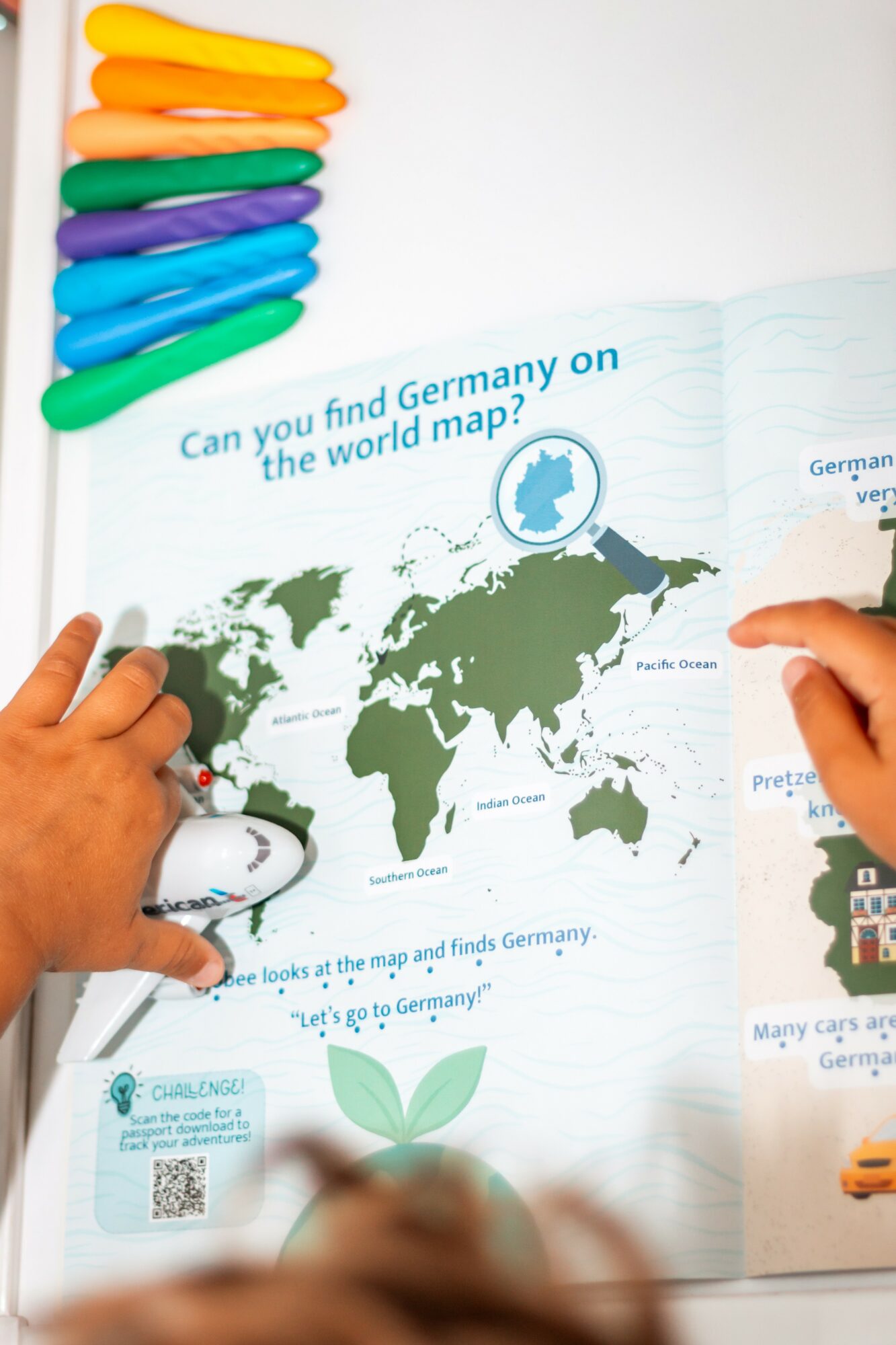 Children's hands point to a world map with a question about Germany and a globe icon, colorful markers at top left.