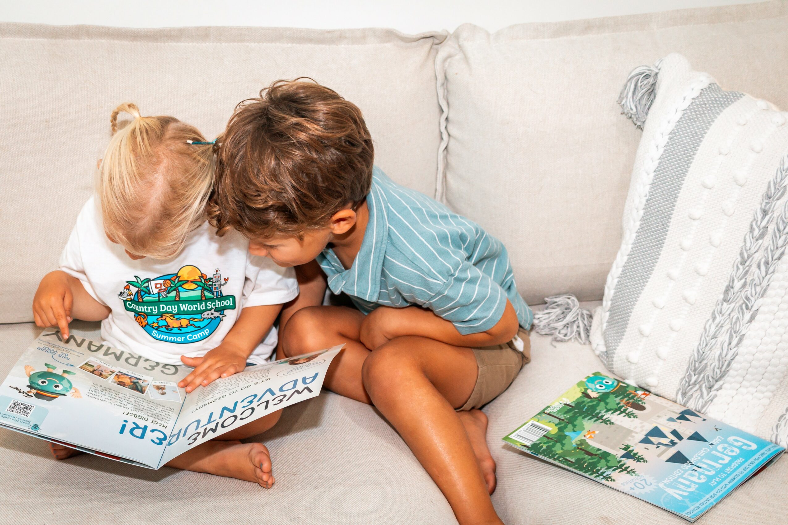 Two children, a girl and a boy, sitting on a couch and reading a book together, with pillows nearby.