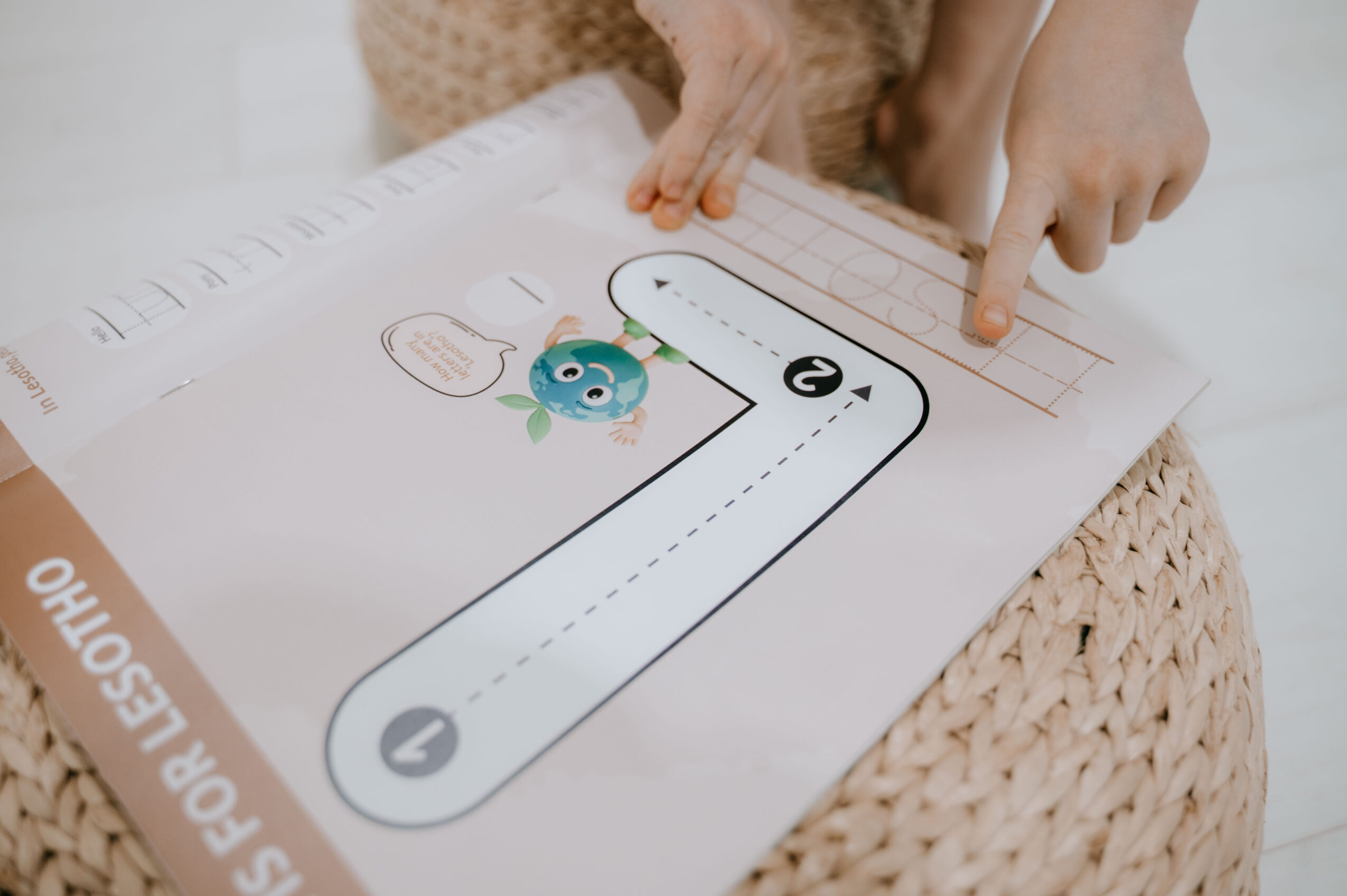 Child's hand points at a worksheet with a cartoon character and a path with numbered circles, on a woven surface.