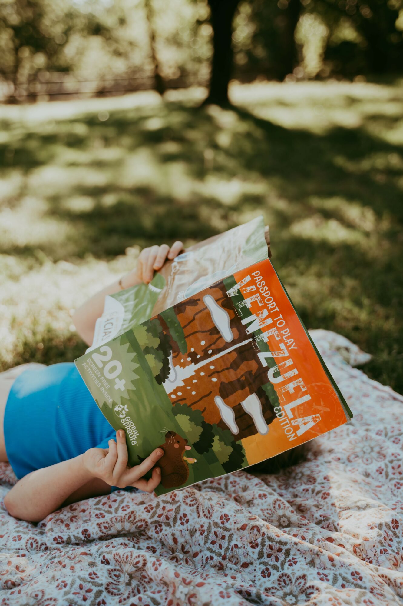 Child lying on a blanket reading a colorful book outdoors with trees and grass in the background.