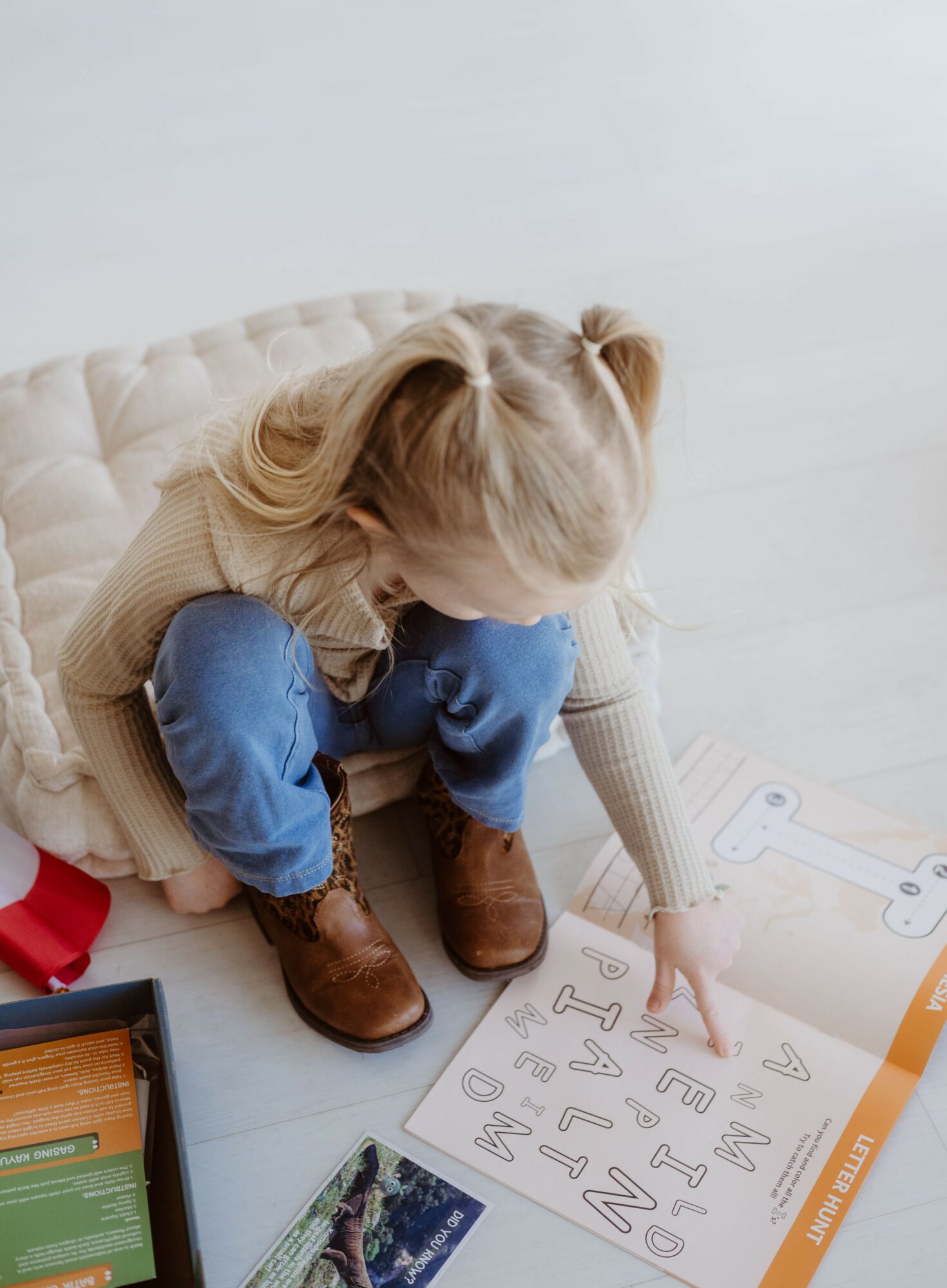 Child with blonde hair, wearing a beige sweater, blue jeans, and brown boots, sitting on a white surface, pointing at a letter chart.