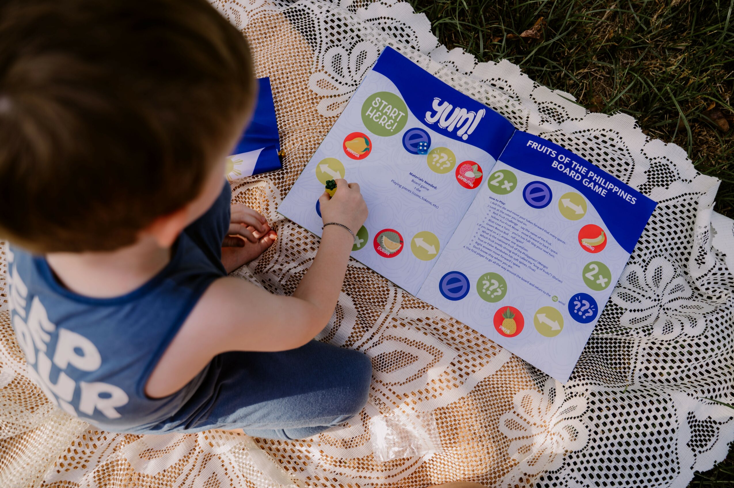 Child with brown hair playing a board game on a lace cloth outdoors, seen from above, with game pieces and cards visible.