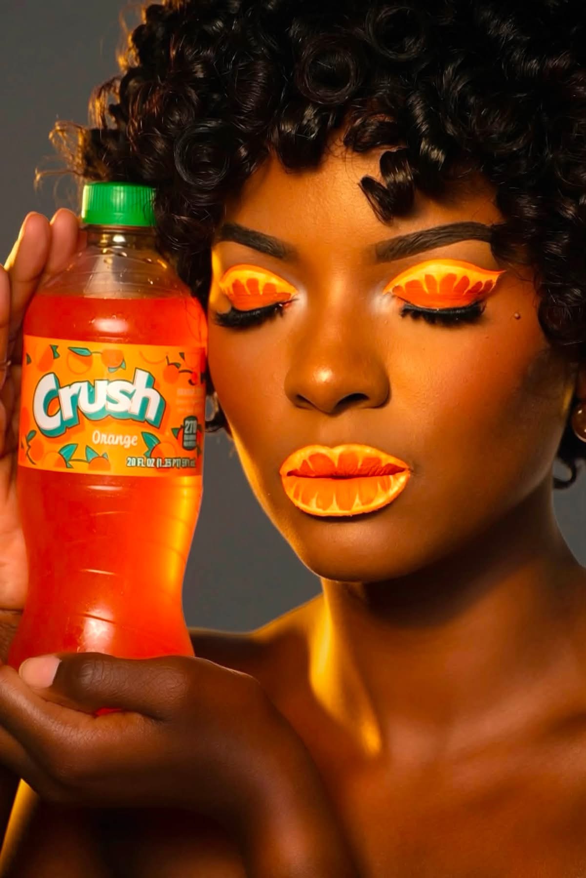 Woman with curly hair and colorful makeup holding a Crush soda bottle, eyes closed, lips painted orange.