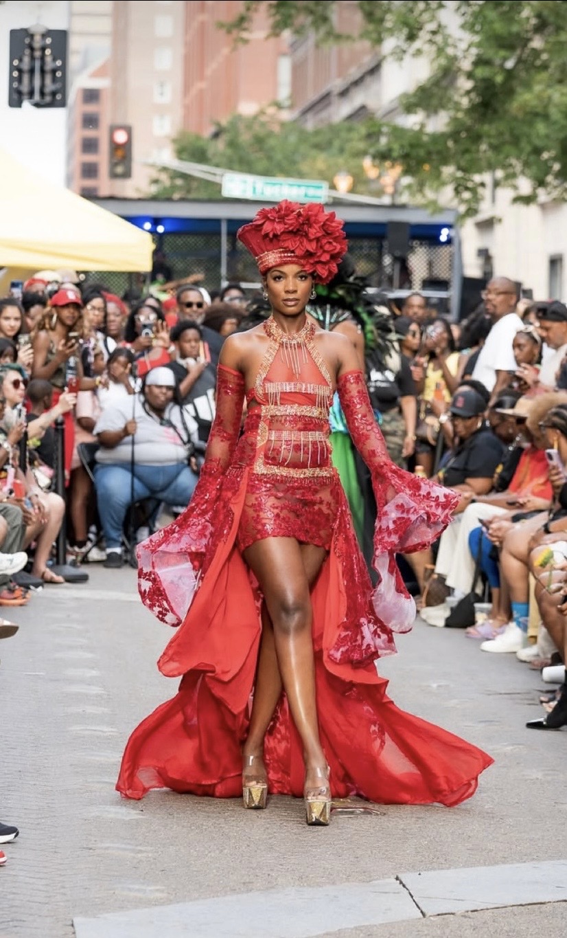 Woman walking in red outfit with a large floral headpiece, surrounded by a crowd at an outdoor event.