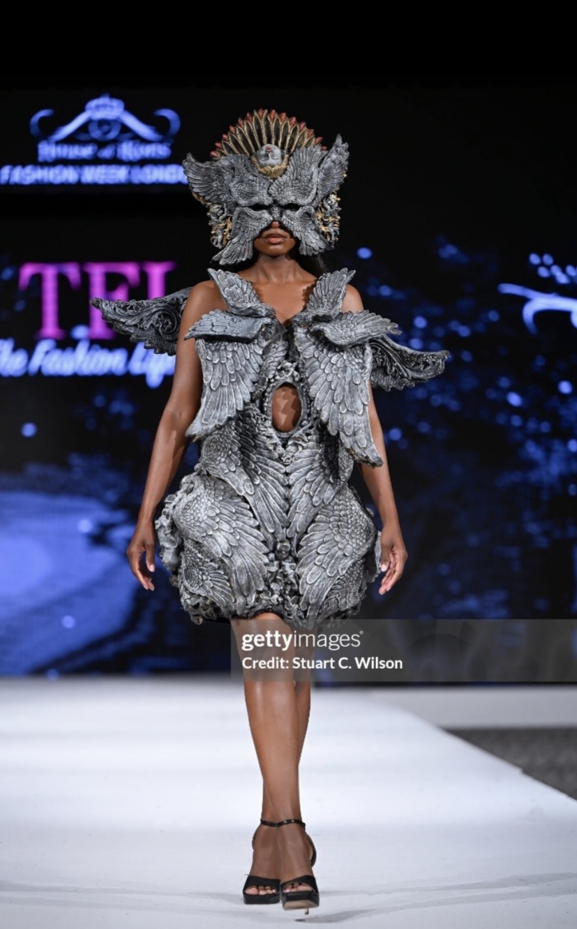 Model walking on runway wearing an elaborate, textured costume with a large headpiece and shoulder details, dark background with text.
