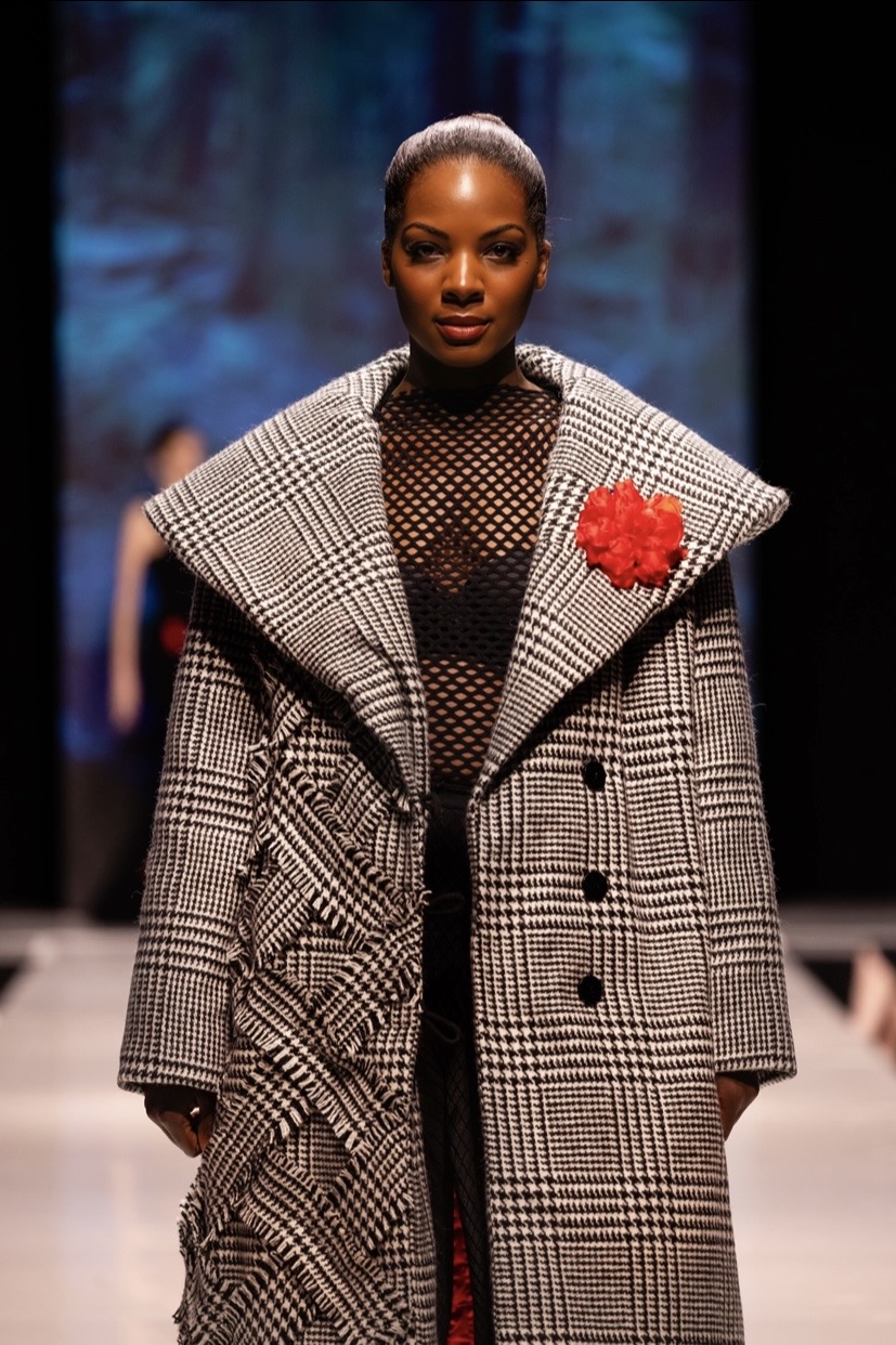 Fashion model walking runway in a checkered coat with a red flower accessory, black mesh top, and dark background.