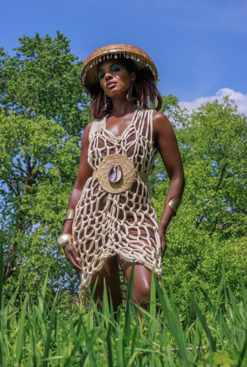 Woman in a crochet dress and straw hat standing outdoors among green grass and trees, under a blue sky.