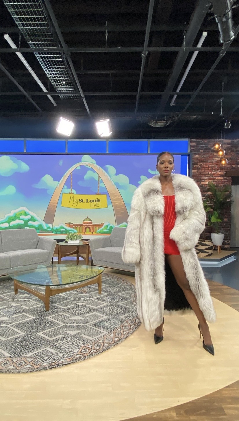 Woman in a red dress and white fur coat standing in a studio with a colorful cartoon background, furniture, and a patterned rug.