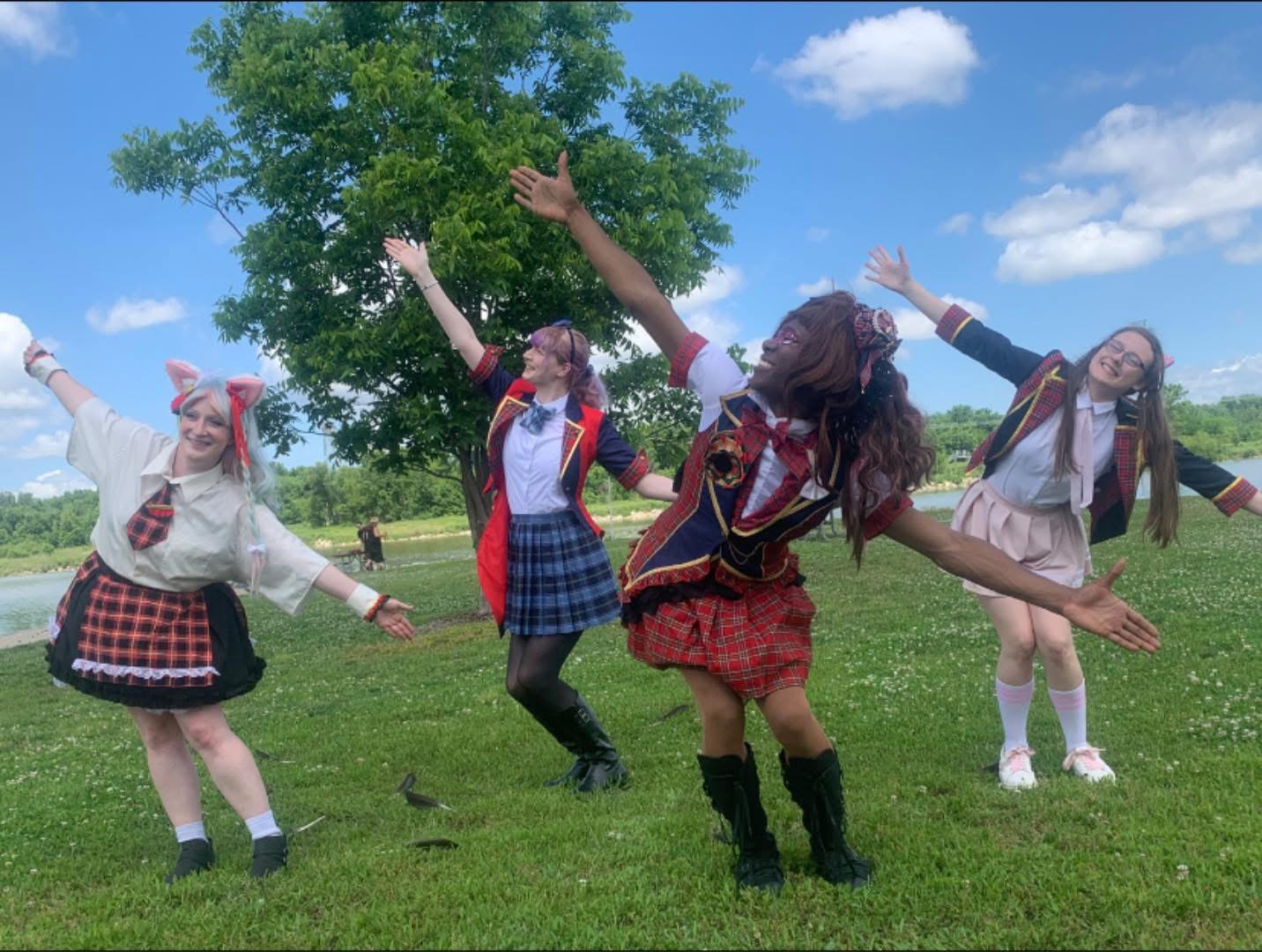 Four girls in school uniforms dancing outdoors on a grassy field with a tree and blue sky.