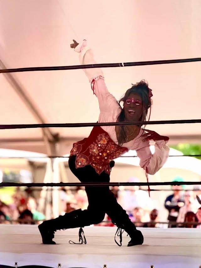 Person in a ring, reaching upward with one arm, wearing a red and pink outfit, with an audience in the background.