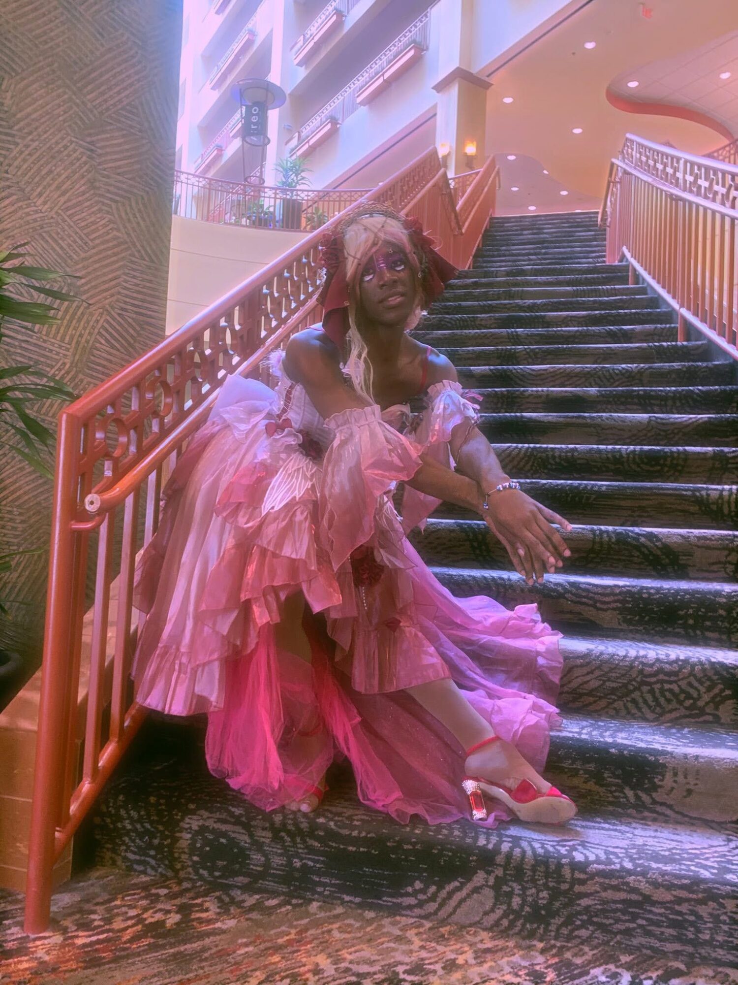 Person in pink dress sitting on staircase with railing, indoors with decorative lighting and plants.