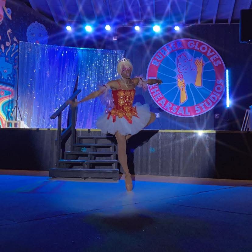 Ballet dancer in costume on stage with blue lighting and a circular logo in the background.