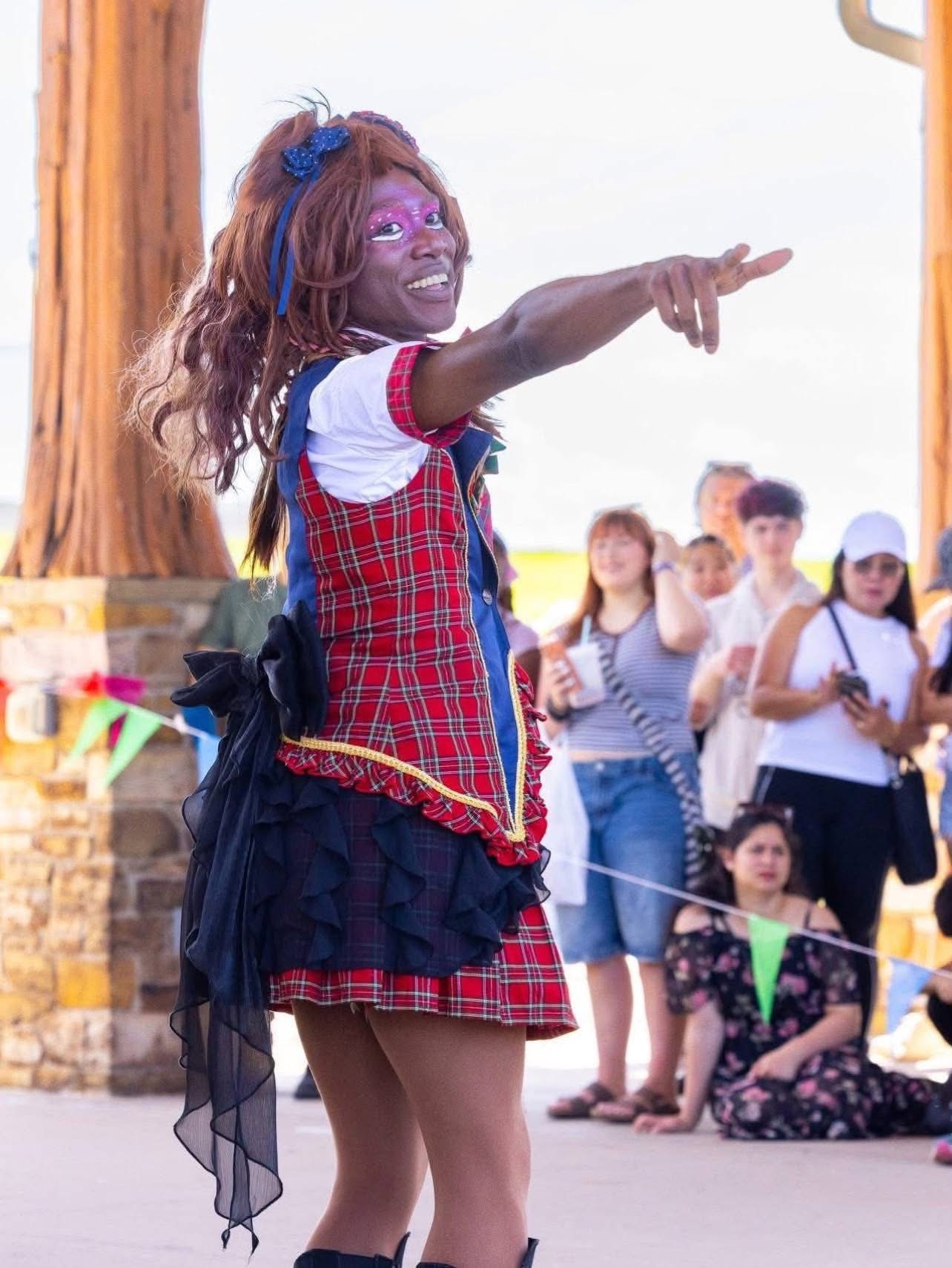 Person dressed as a clown with colorful makeup and costume, pointing and smiling outdoors with a group of children in the background.