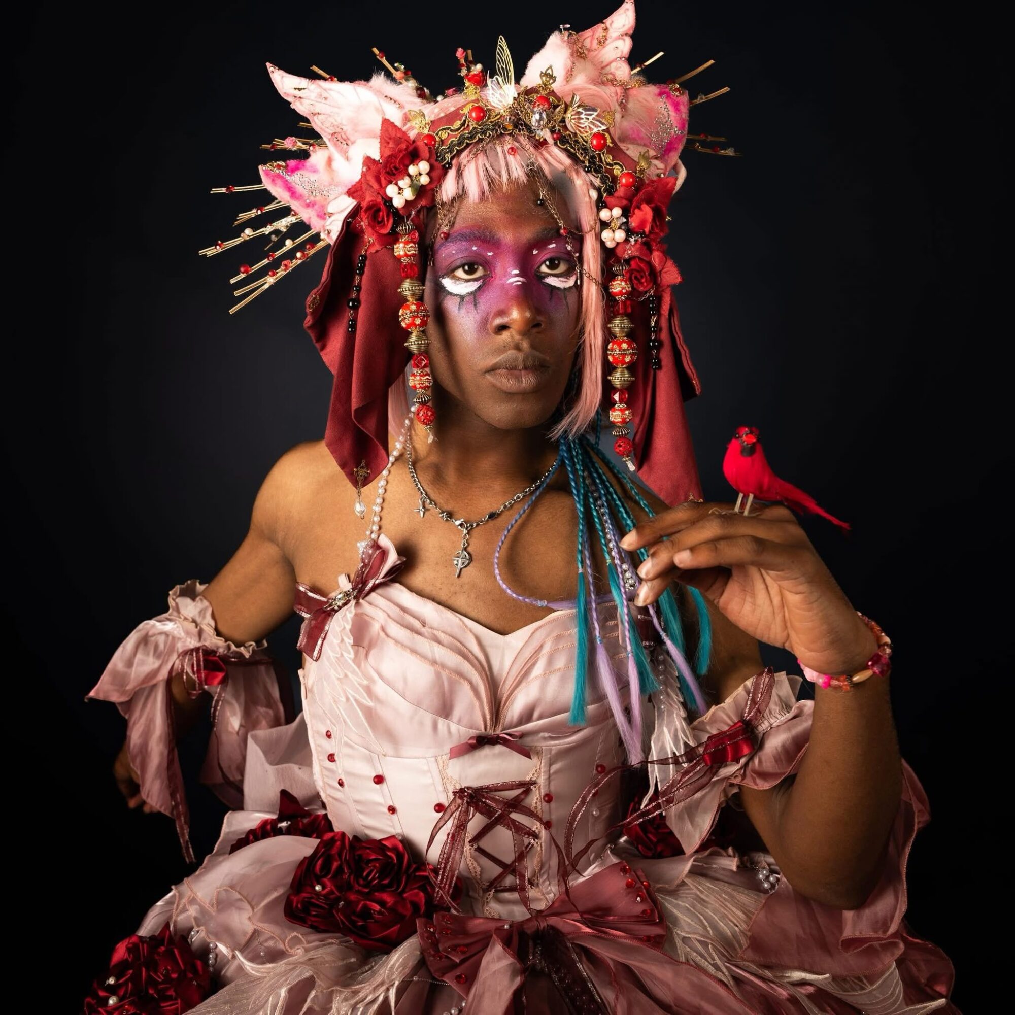 Person with elaborate pink and red costume, headpiece, and colorful makeup holding a red bird on finger, dark background.