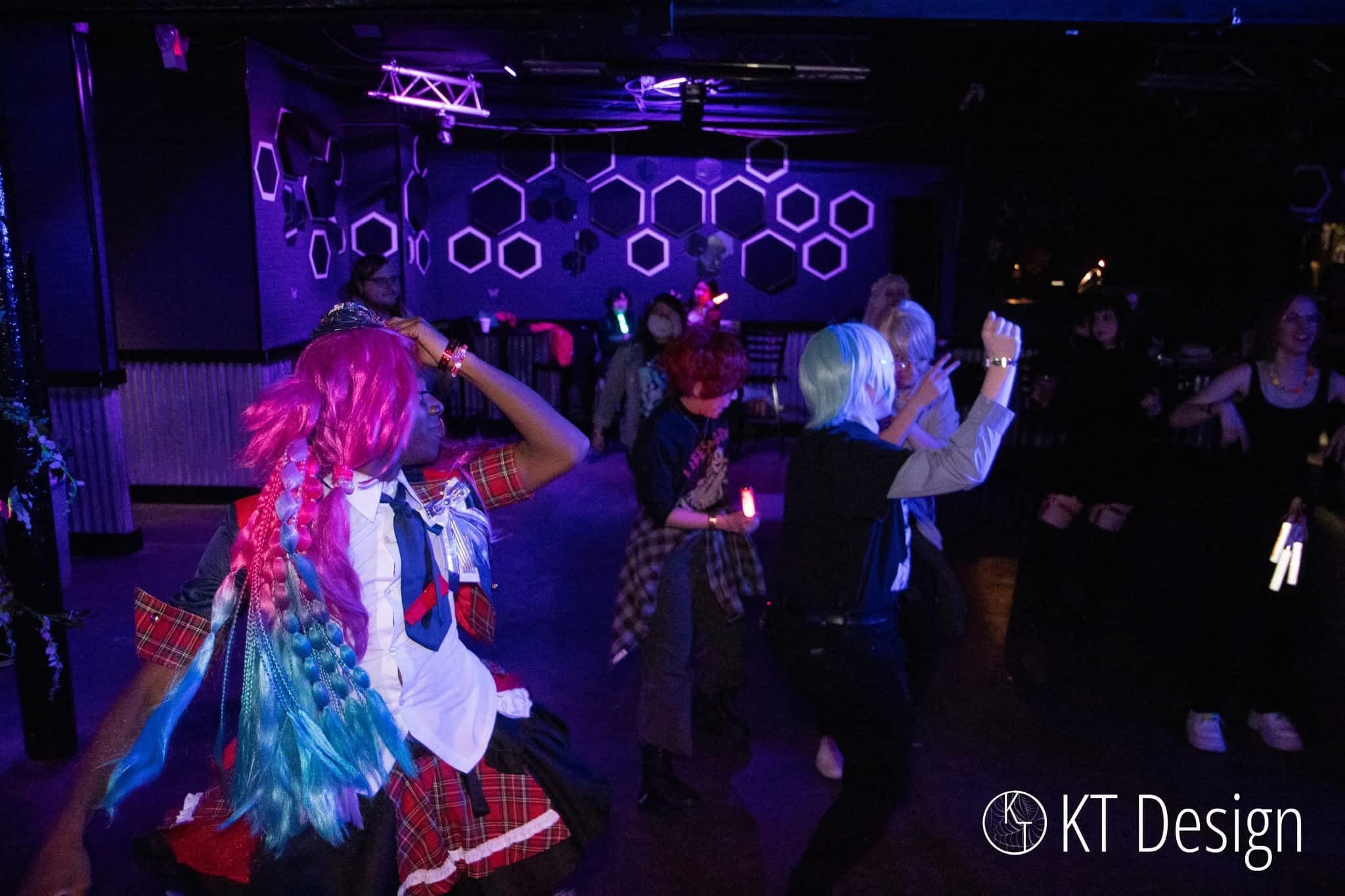 People dancing in a dark room with purple hexagon patterns on the wall, some wearing colorful wigs and costumes.