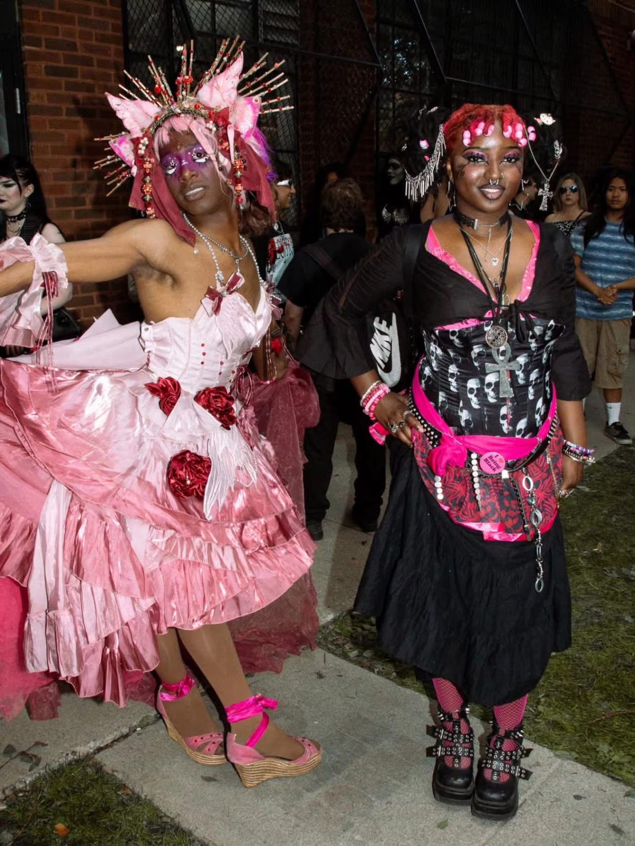 Two women in colorful, elaborate costumes with accessories and makeup, standing outdoors at night with a crowd in the background.