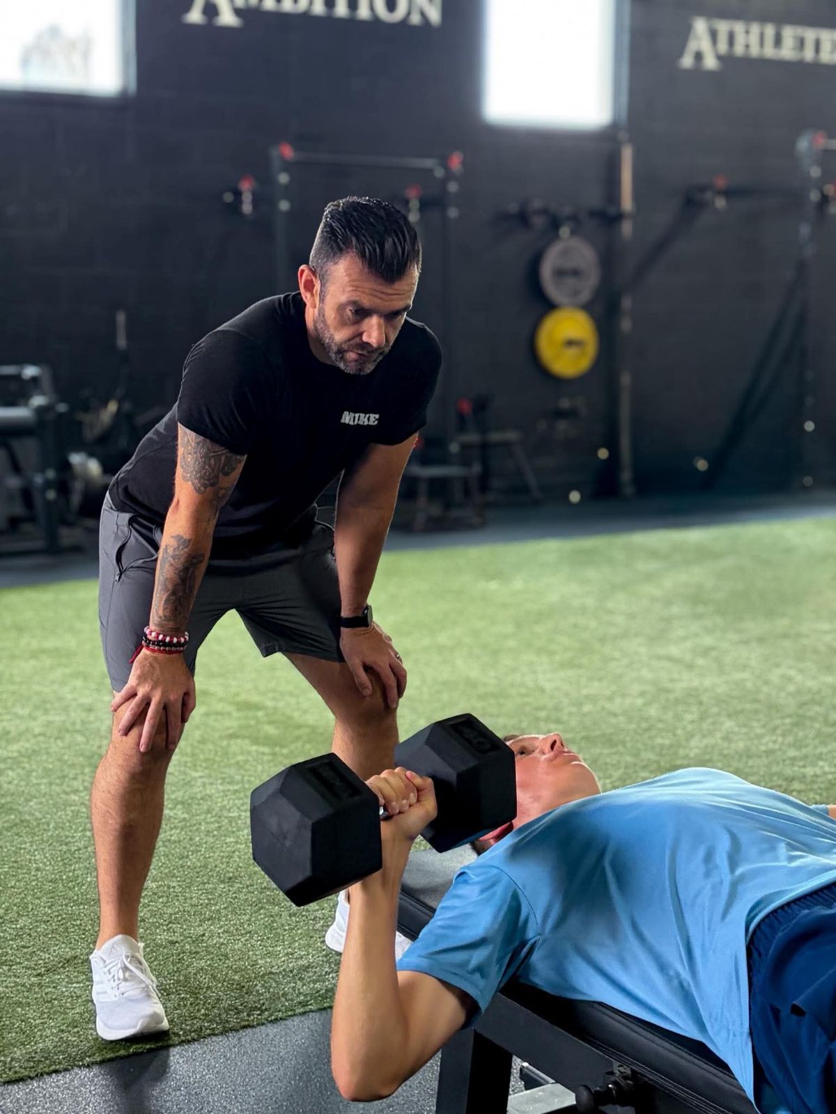 A man in black workout clothes assists a person lying on a bench lifting a dumbbell in a gym. The gym has black walls and equipment.