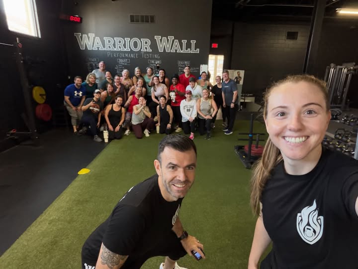 Group of people in a gym, some kneeling and some standing, with a woman taking a selfie in the foreground, smiling.