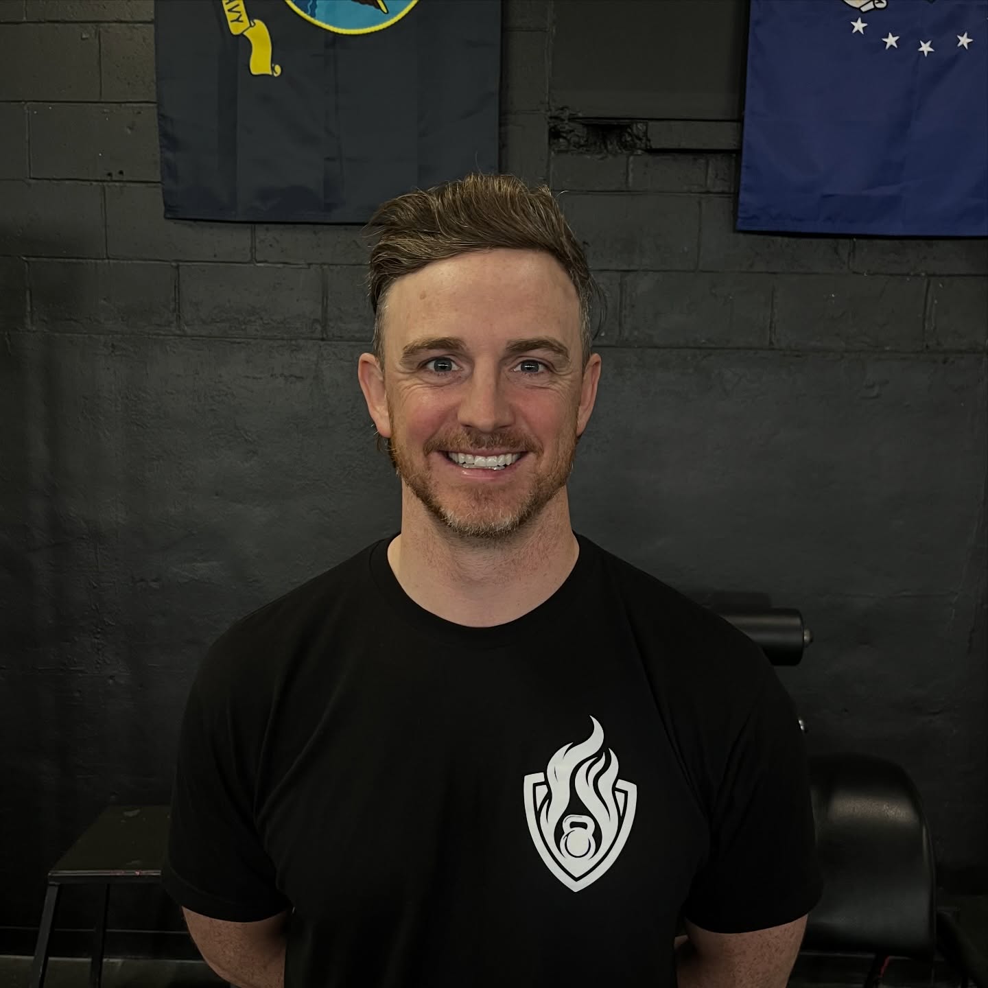 Smiling man with short brown hair and beard, wearing a black T-shirt with a white flame logo, standing indoors.