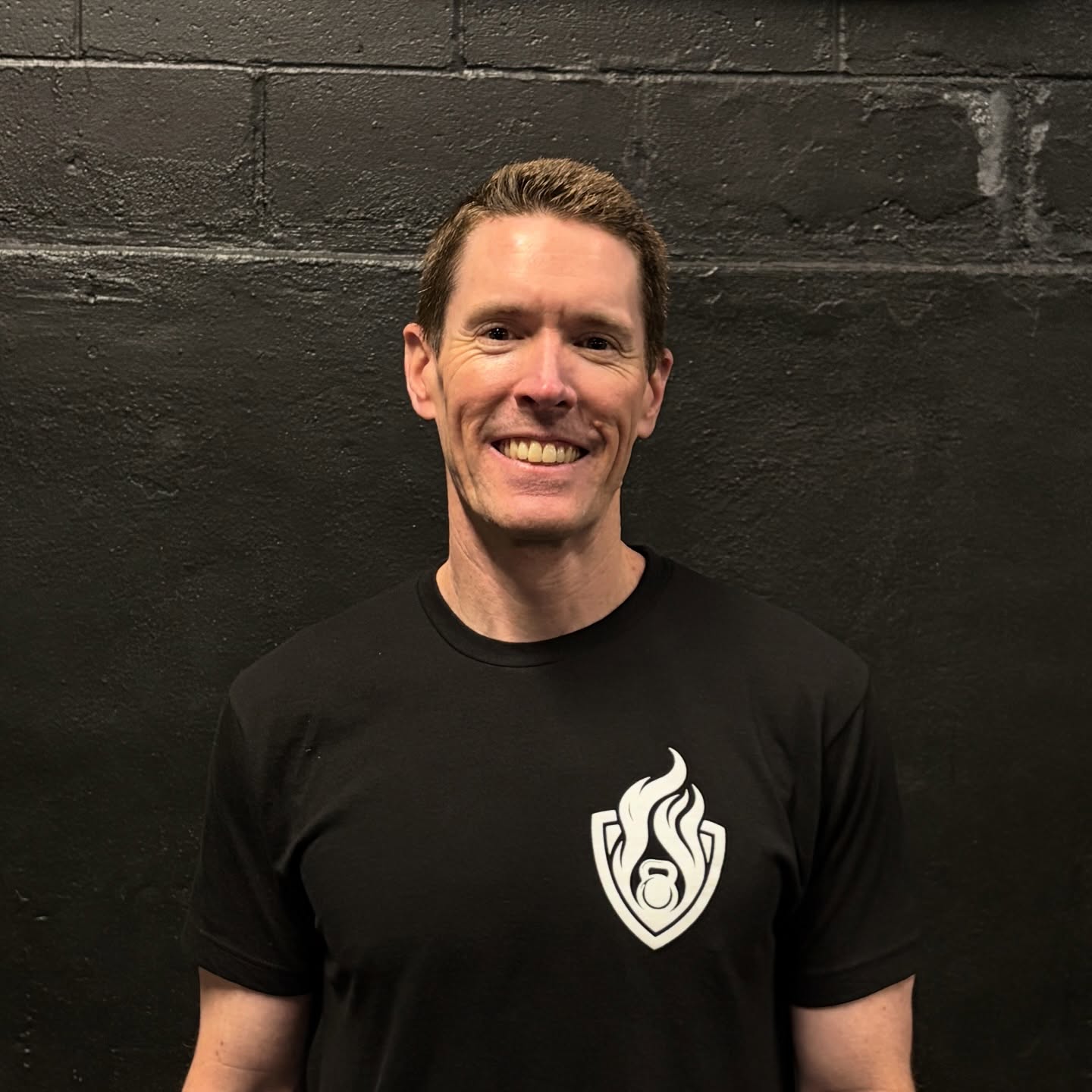 Smiling man with short hair wearing a black T-shirt with a white flame logo, standing against a dark wall.