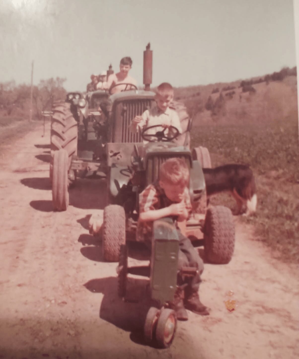 Children riding small tractors on a dirt road in a rural area, with open fields in the background.