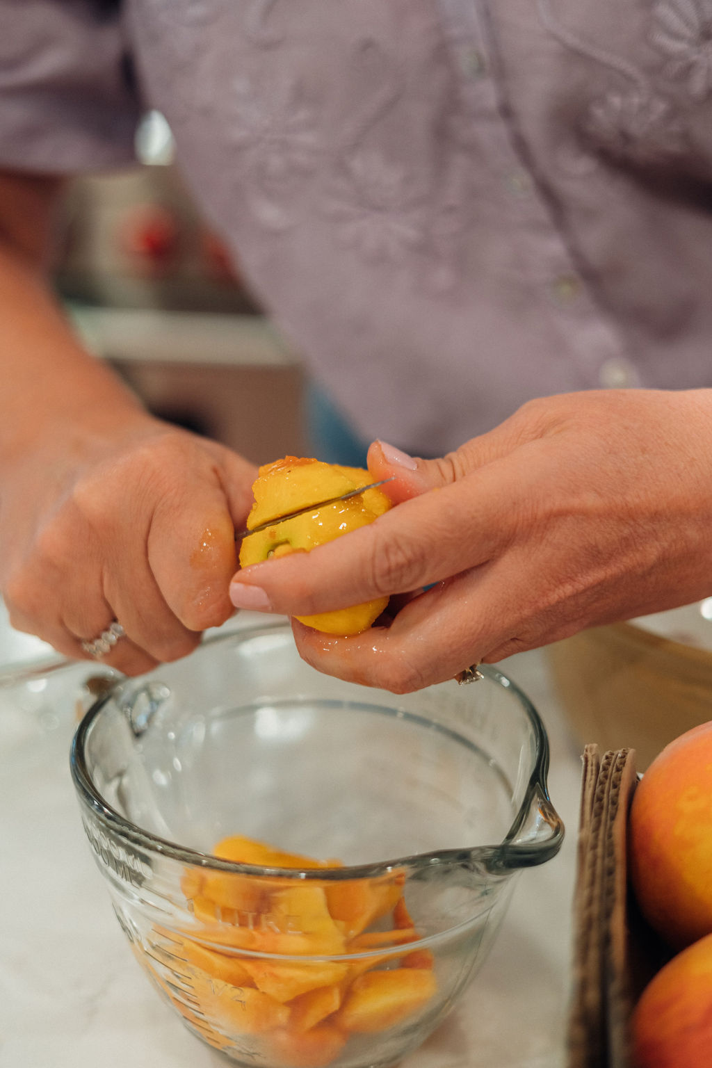 Two people squeeze a yellow fruit, possibly a lemon or orange, into a glass bowl with sliced fruit inside.