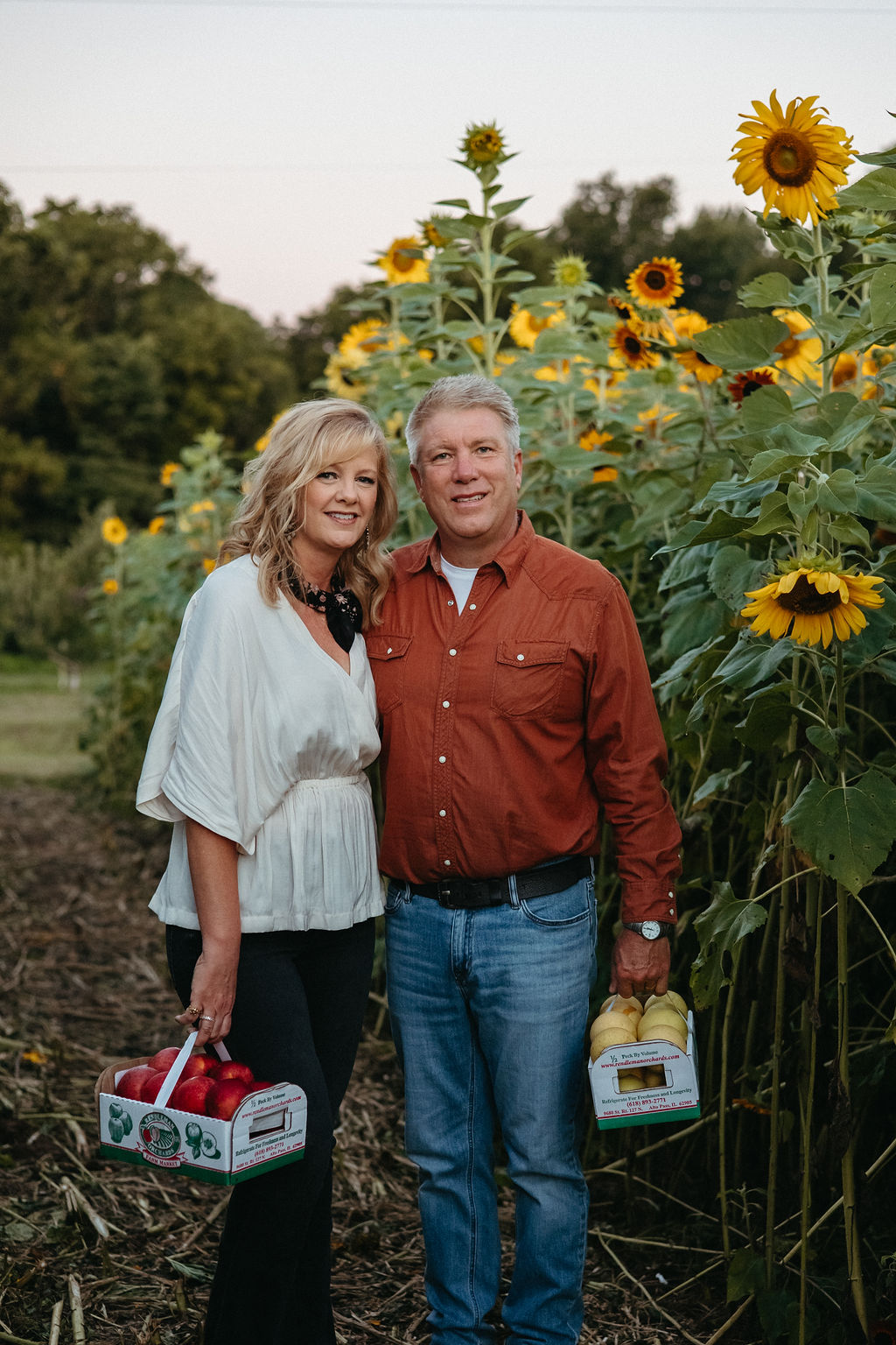 A smiling couple stands among tall sunflowers outdoors, holding picnic baskets, with trees and a cloudy sky in the background.