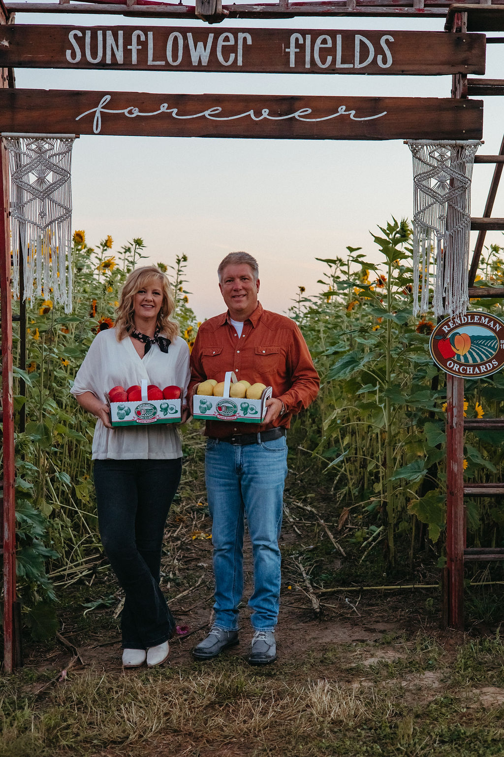 A man and woman stand in a flower field holding boxes, under a sign that reads 'Sunflower Fields Forever'.