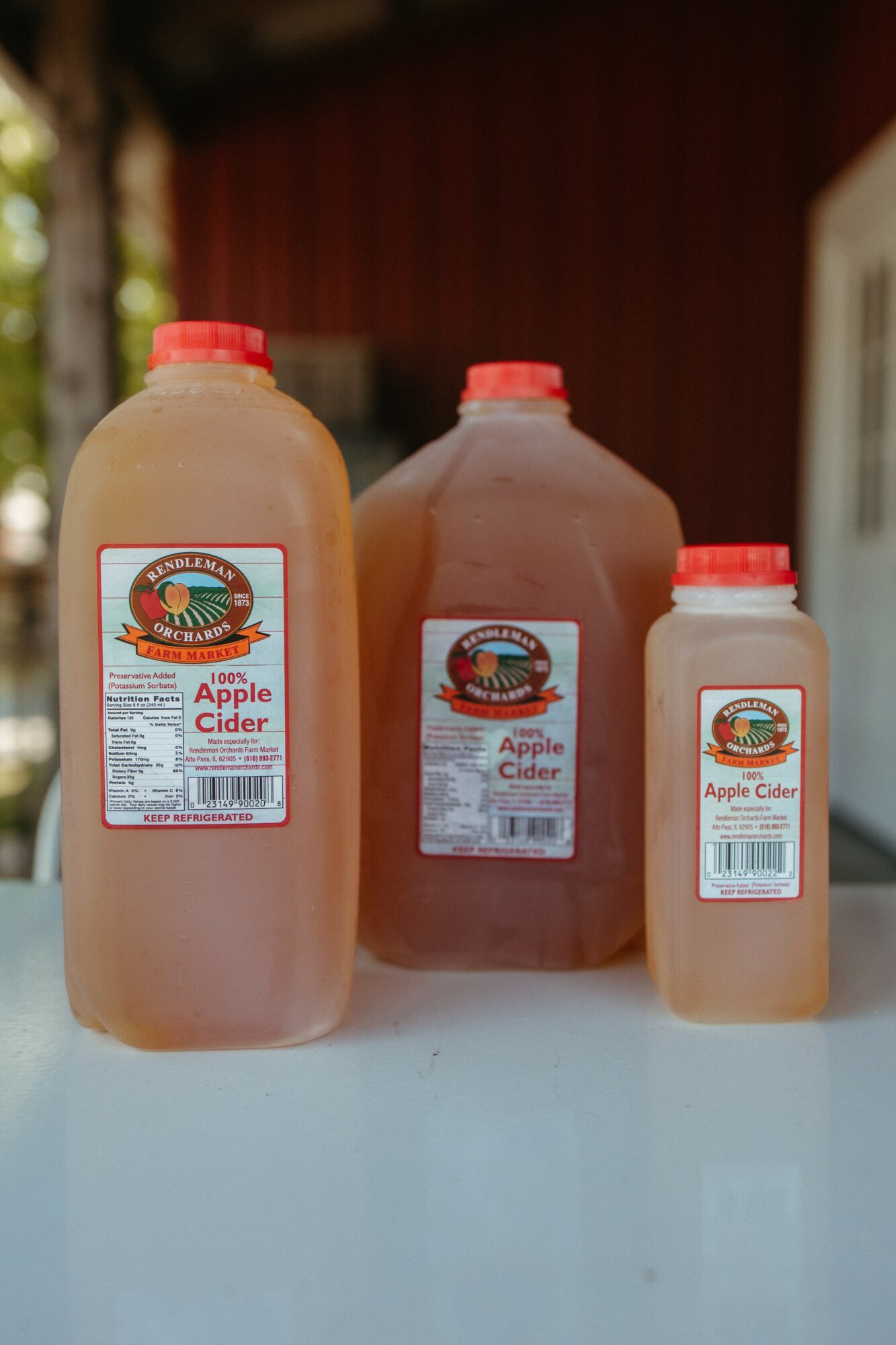 Three bottles of apple cider on a white surface, with a red and brown background and trees visible in the distance.
