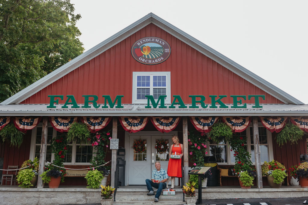 Red barn-style building with a sign reading 'FARM MARKET' and decorative hanging plants, flowers, and a person sitting outside.