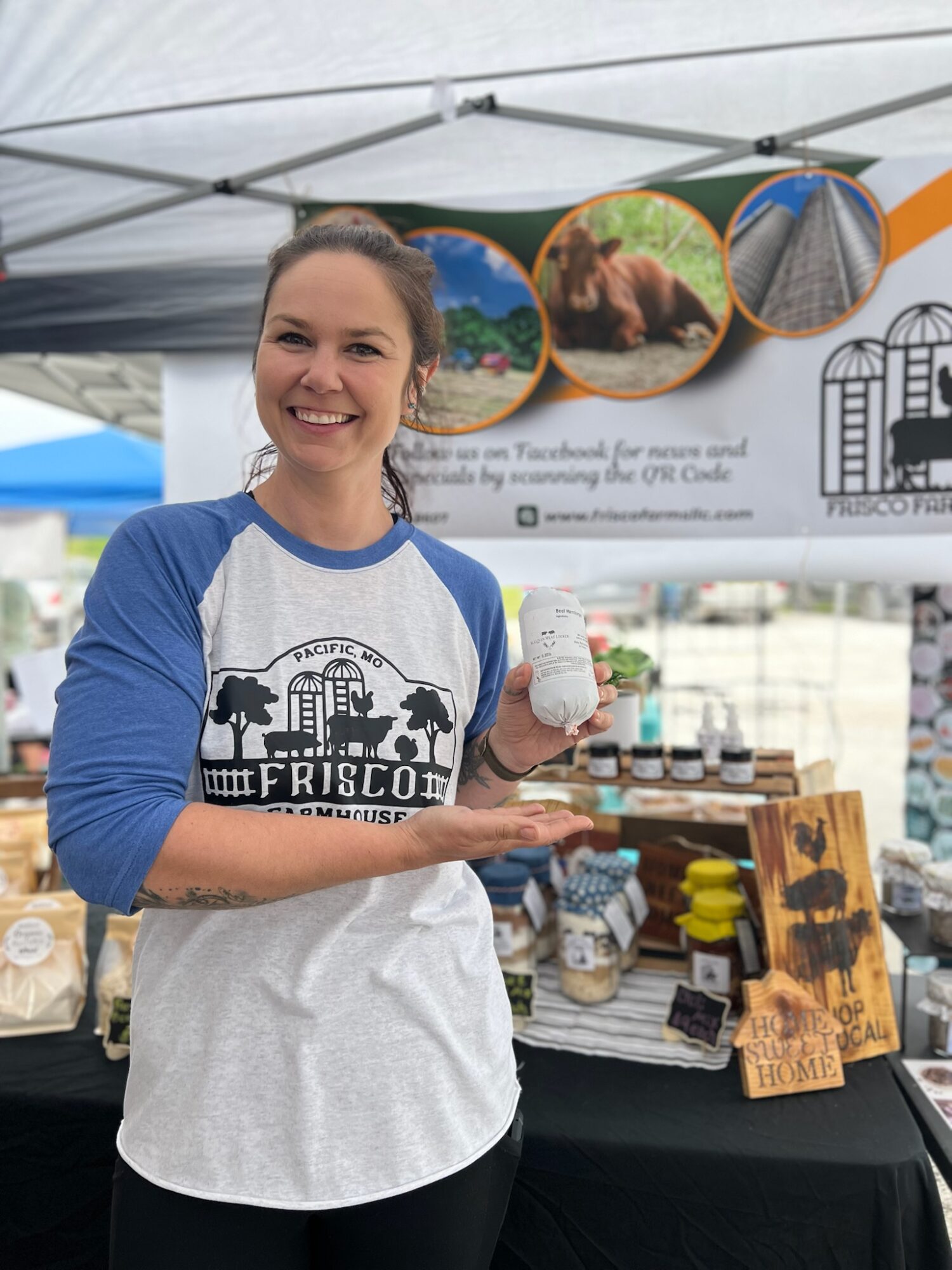 Smiling woman holding a small white object at an outdoor market stall with framed pictures and products displayed.