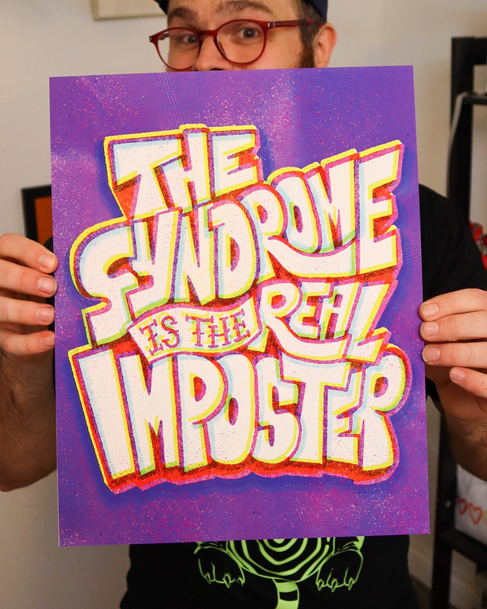Person holding a colorful sign with bold text, partially visible face, glasses, and a dark shirt, background indoors.