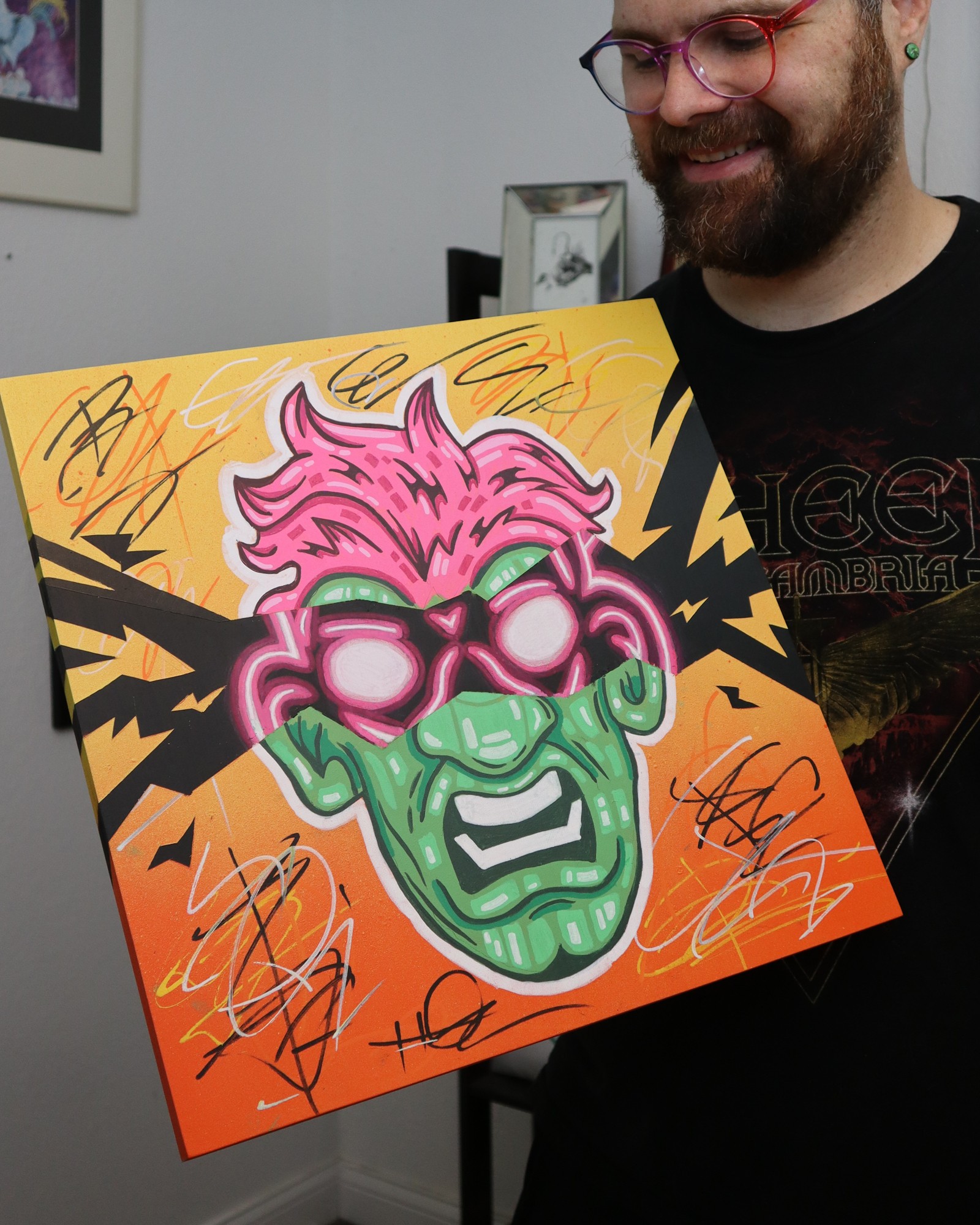 Person holding colorful graffiti-style artwork of a green face with pink hair and sunglasses, smiling, with a yellow and orange background.