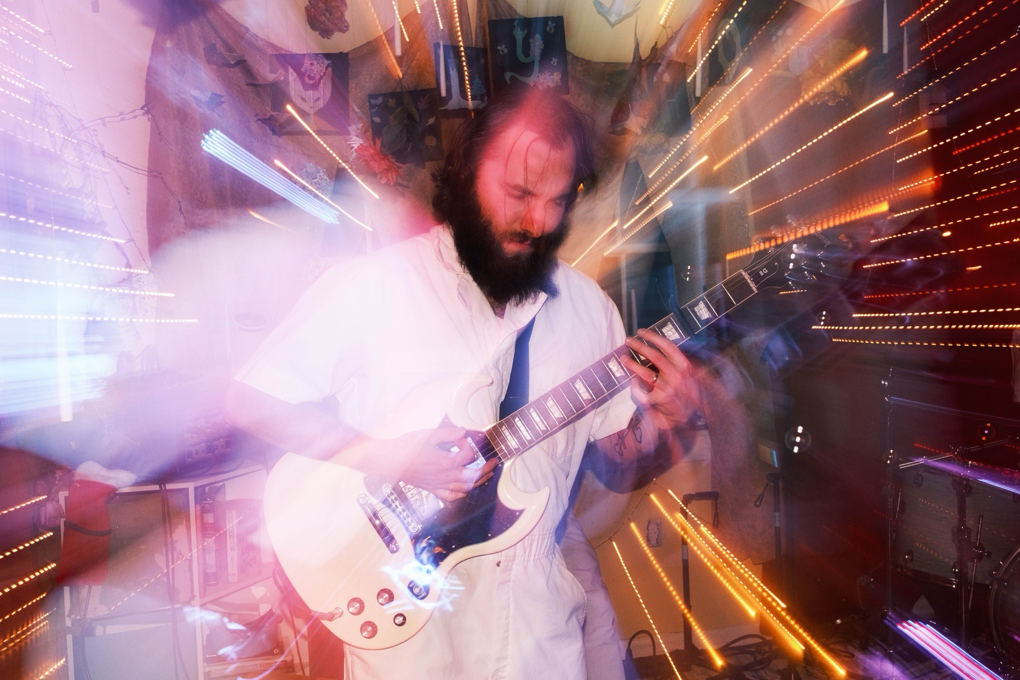 Person playing guitar with colorful light streaks in background, motion blur effect, white shirt, beard, and curly hair.