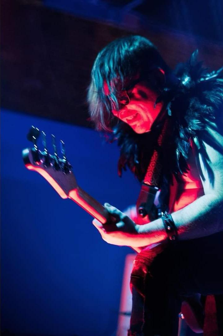 Person with dark hair playing guitar on stage, wearing feathered clothing, illuminated by colorful stage lights.