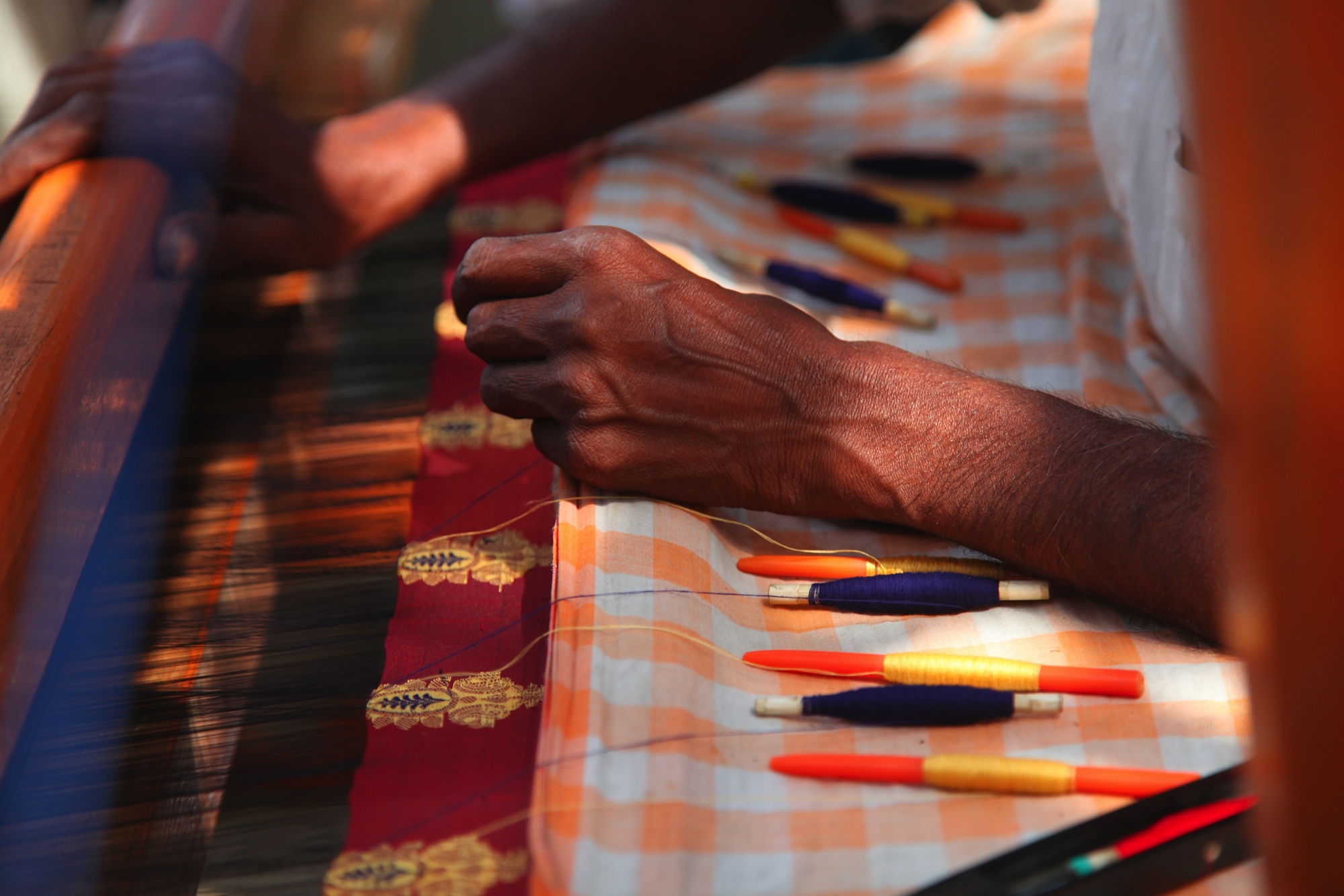 Close-up of hands working on a textile loom with colorful threads and tools, fabric with checkered pattern visible.