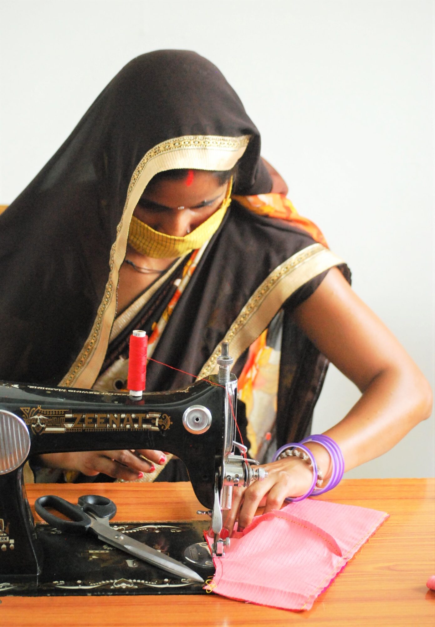 Woman sewing fabric with a sewing machine, wearing traditional clothing and jewelry, on a wooden table.