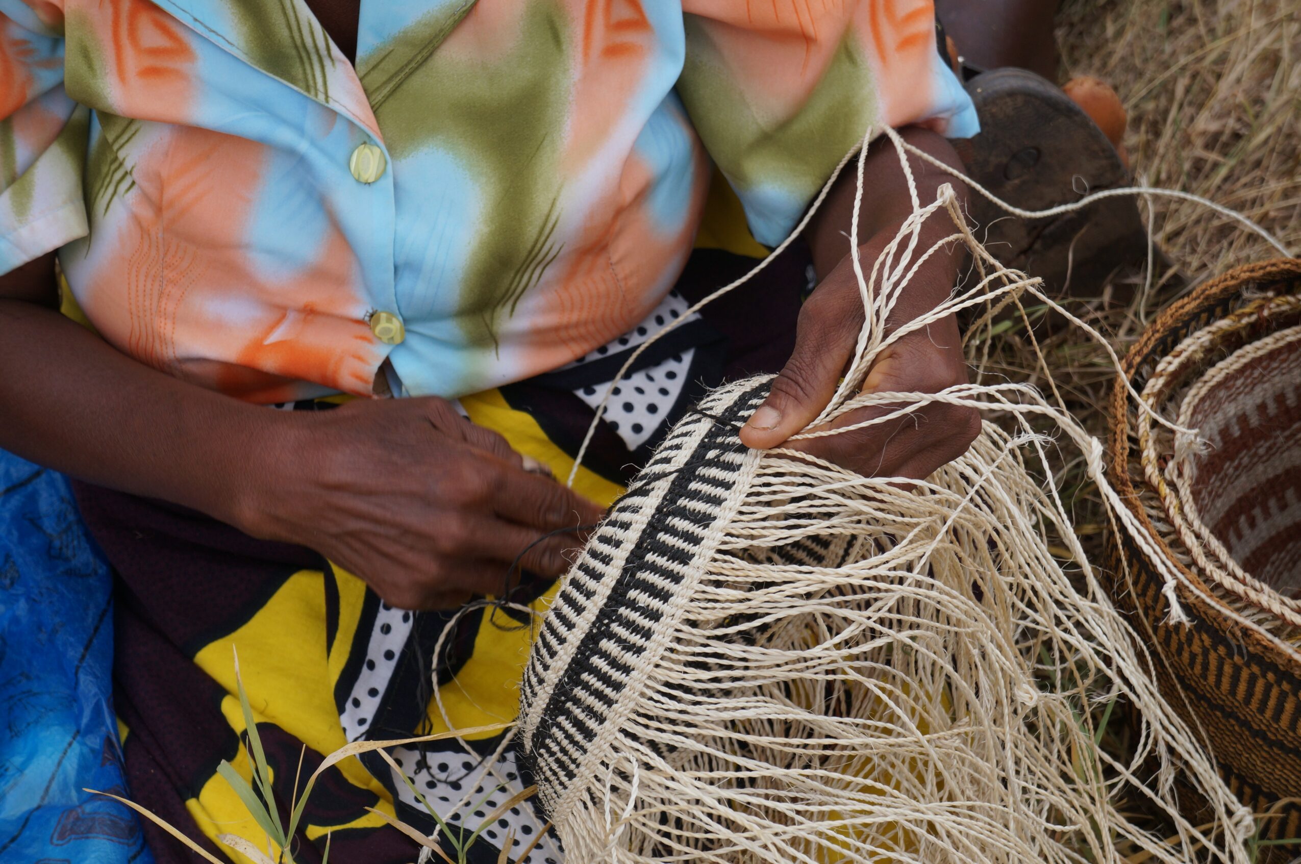 Person weaving with straw, wearing colorful clothing, surrounded by woven baskets and straw materials.
