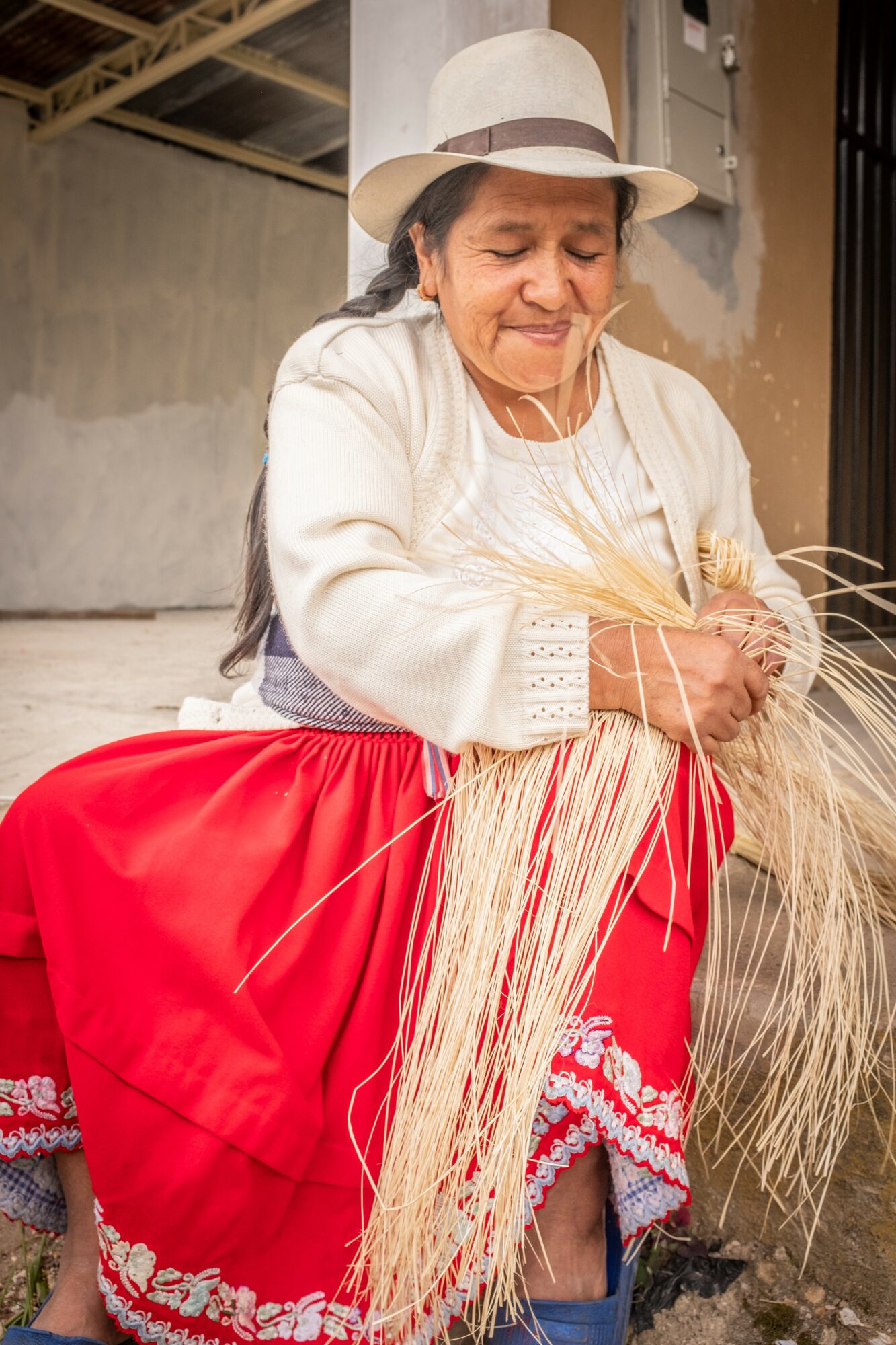 Elderly woman in traditional clothing, wearing a white hat, sitting outdoors, holding straw, with a neutral expression.