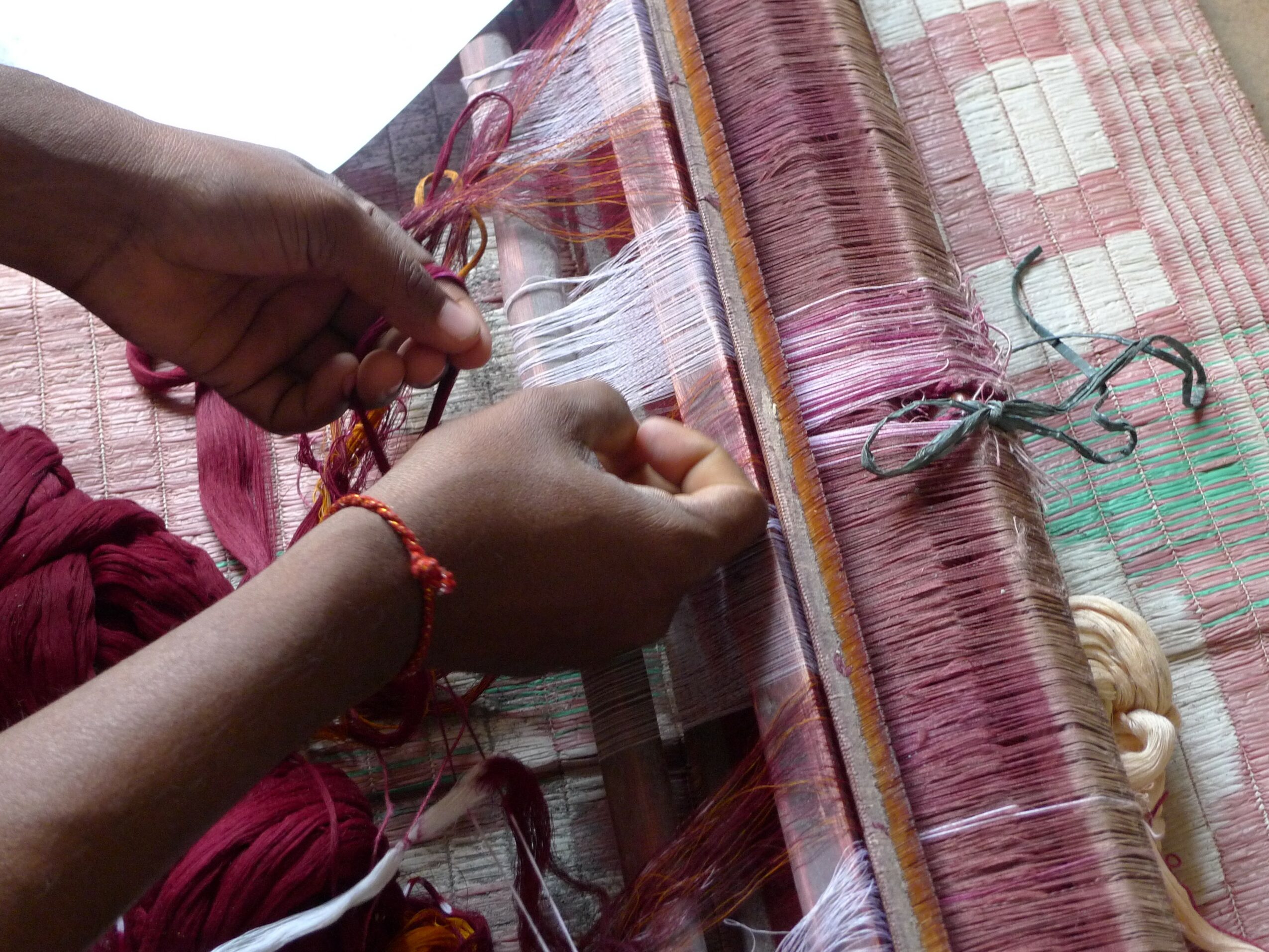 Person weaving colorful threads on a loom, hands working with yarn and fabric strips, pink and purple hues visible.
