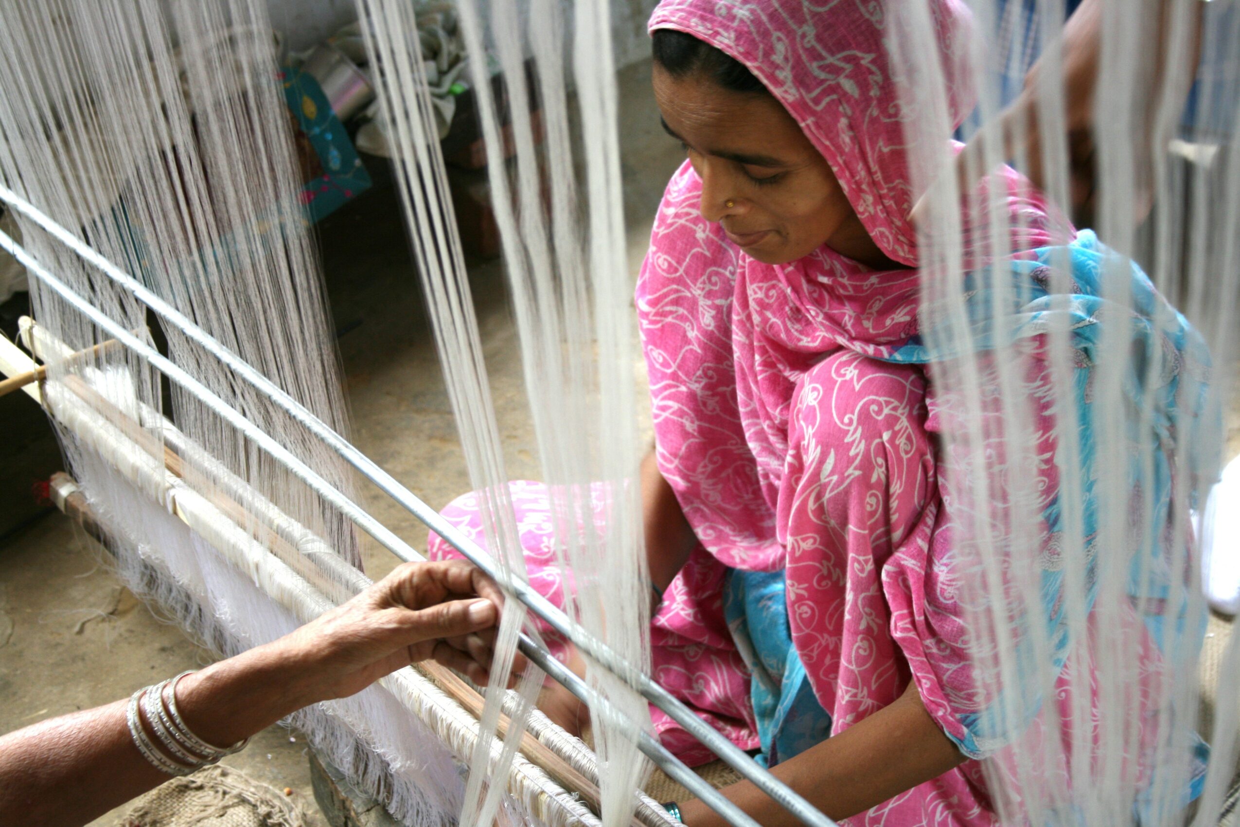 Girl in pink traditional clothing weaving fabric on a loom, with a person assisting nearby.