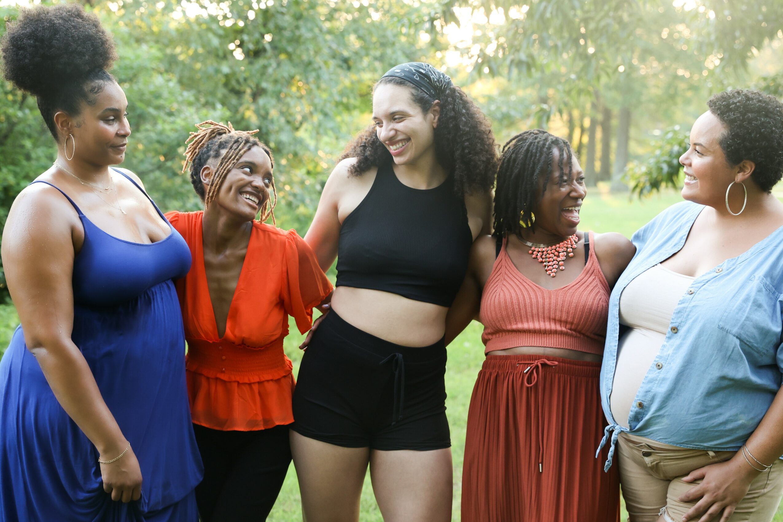 Group of five women smiling and talking outdoors in a park with trees and sunlight.