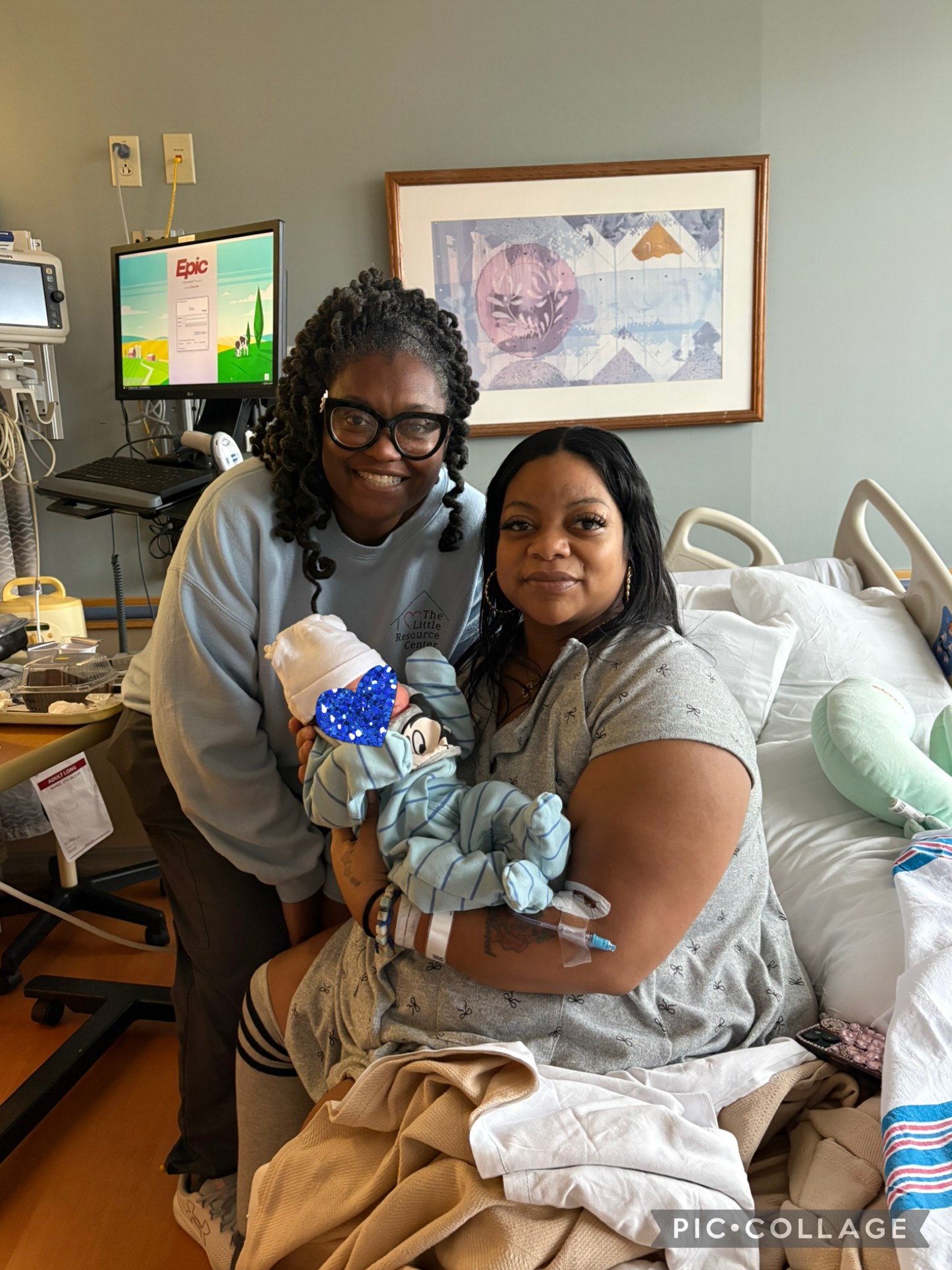 Two women in a hospital room, one holding a newborn baby, smiling at the camera, with medical equipment and artwork in the background.