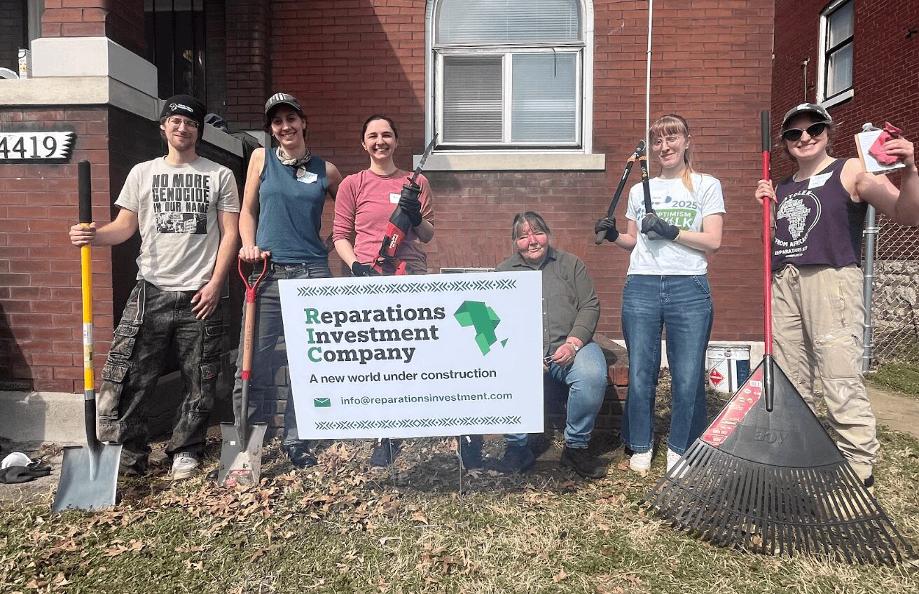 Group of six people holding a sign in front of a brick building, some with tools and rakes, smiling outdoors.